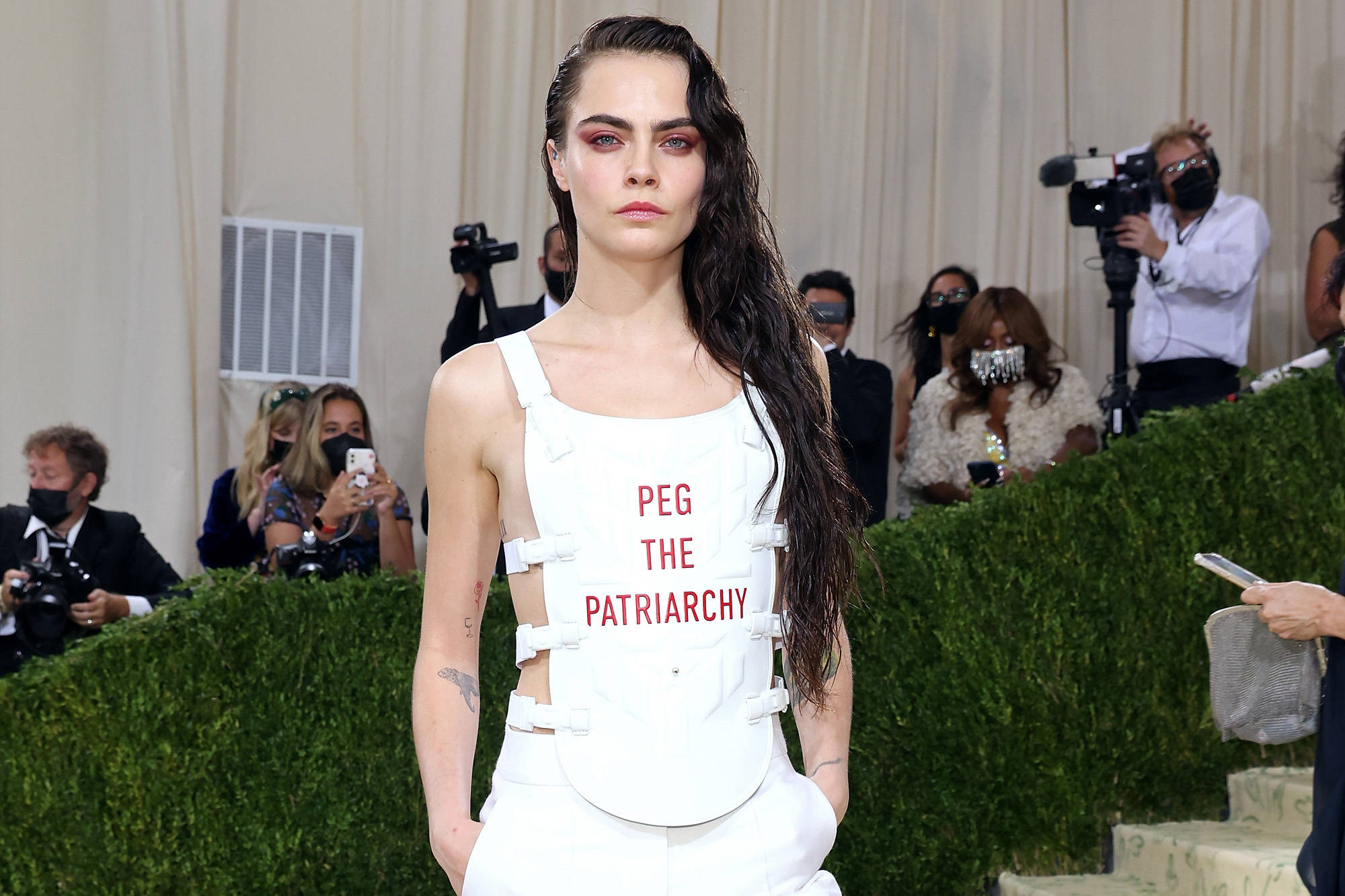 Cara 桌布 壁紙 — 模特兒Cara Delevingne穿著白色束身服裝，上面有紅色文字寫著「PEG THE PATRIARCHY」，在紅地毯上行走，攝影師們捕捉她的身影。
