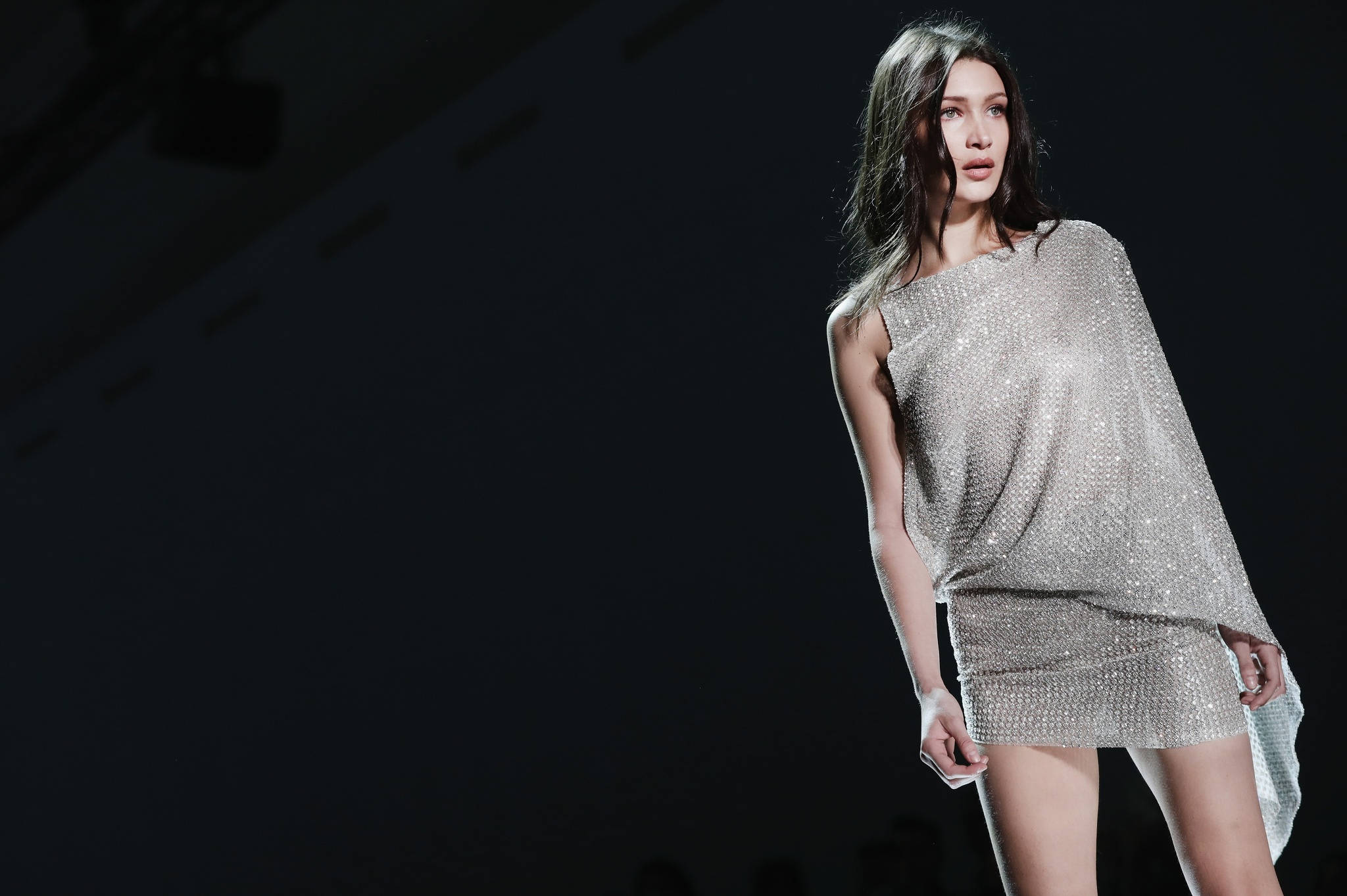 Bella Hadid 桌布 壁紙 — 模特兒在戲劇性的伸展台燈光下，自信地穿著閃閃發光的銀色亮片連衣裙行走。