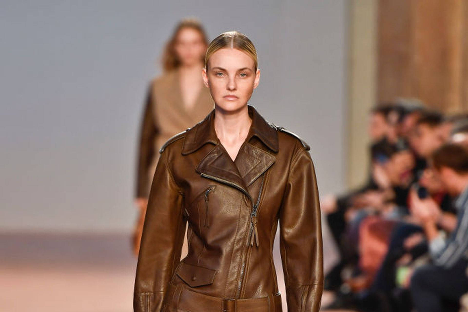 Ferragamo 桌布 壁紙 — 模特兒在明亮的時裝秀燈光下，穿著結構感十足的棕色皮夾克自信地走在伸展台上。