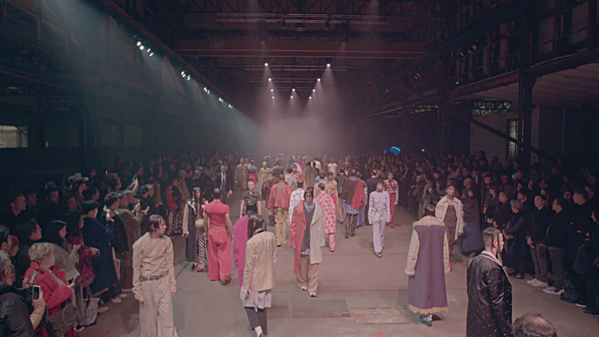 BTS 桌布 壁紙 — 模特兒在時裝秀中，身著色彩繽紛的服裝，在昏暗的工業倉庫中霧氣瀰漫的伸展台上遊行。