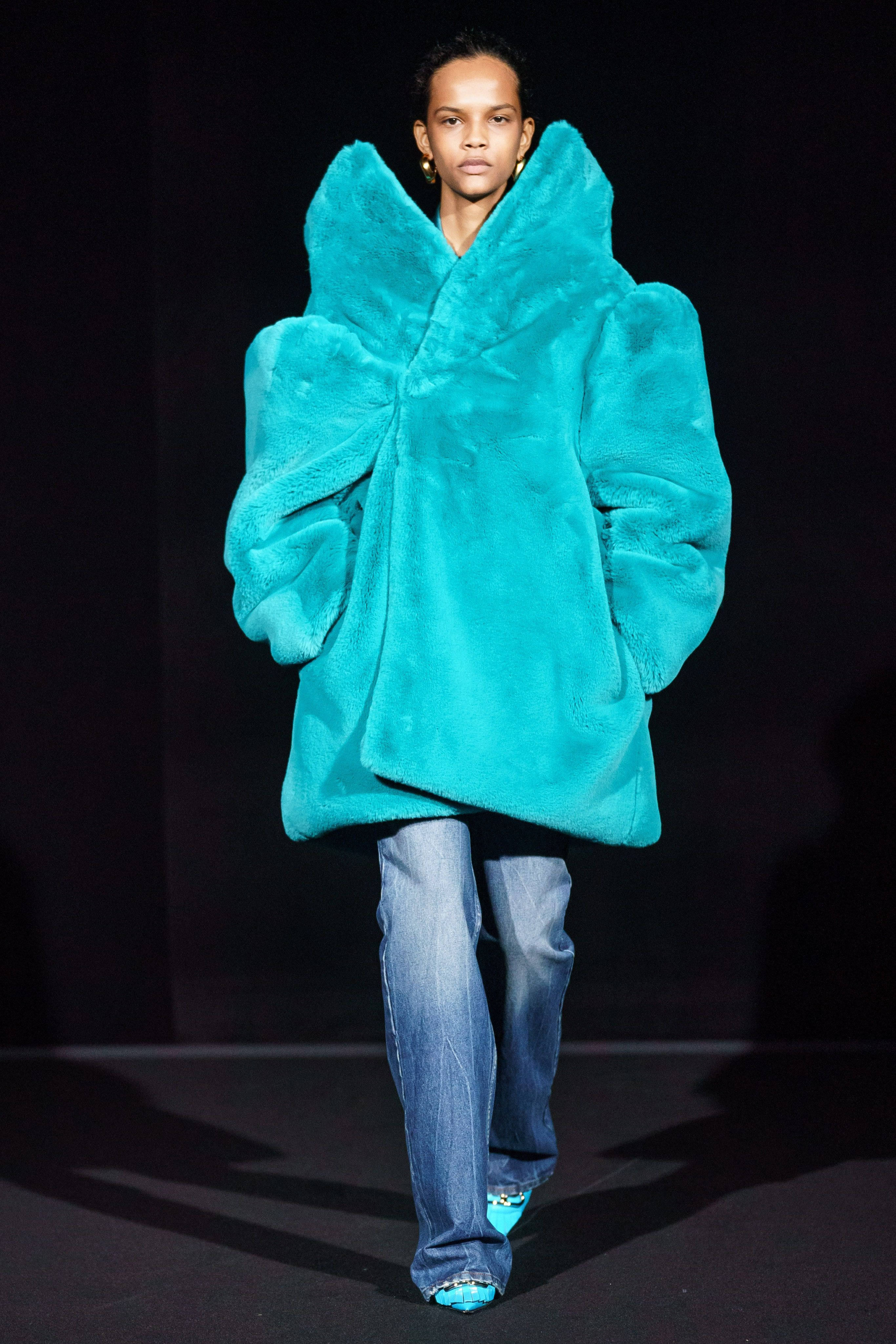 Balenciaga 桌布 壁紙 — 模特兒在舞台燈光下自信地穿著一件寬大的青綠色人造毛皮大衣和藍色牛仔褲。