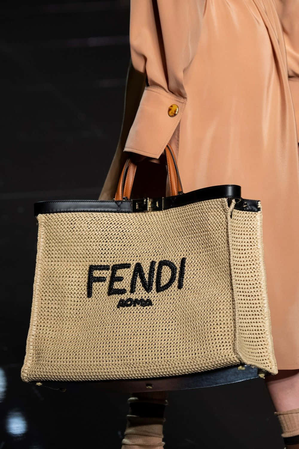 Fendi 桌布 壁紙 — 模特兒手持一款編織FENDI ROMA手提袋，搭配黑色皮革飾邊和手柄，背景為昏暗的時裝伸展台。