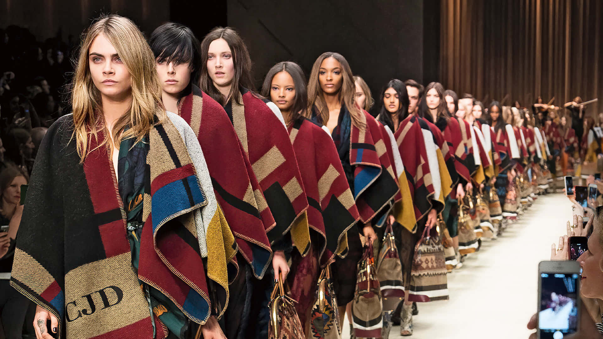 Burberry 桌布 壁紙 — 模特兒披著大膽幾何圖案毛毯，手持相配手袋，在明亮燈光下走過天橋。