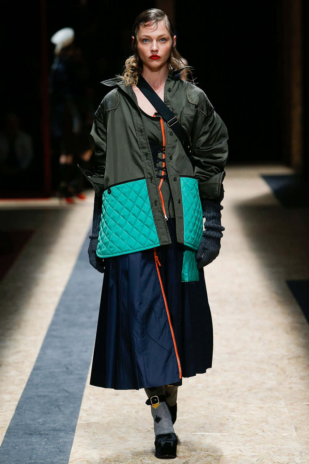 Prada 桌布 壁紙 — 模特兒穿著大膽的綠色絎縫外套，搭配青綠色拼接和橙色拉鍊細節，走在時裝展台上。