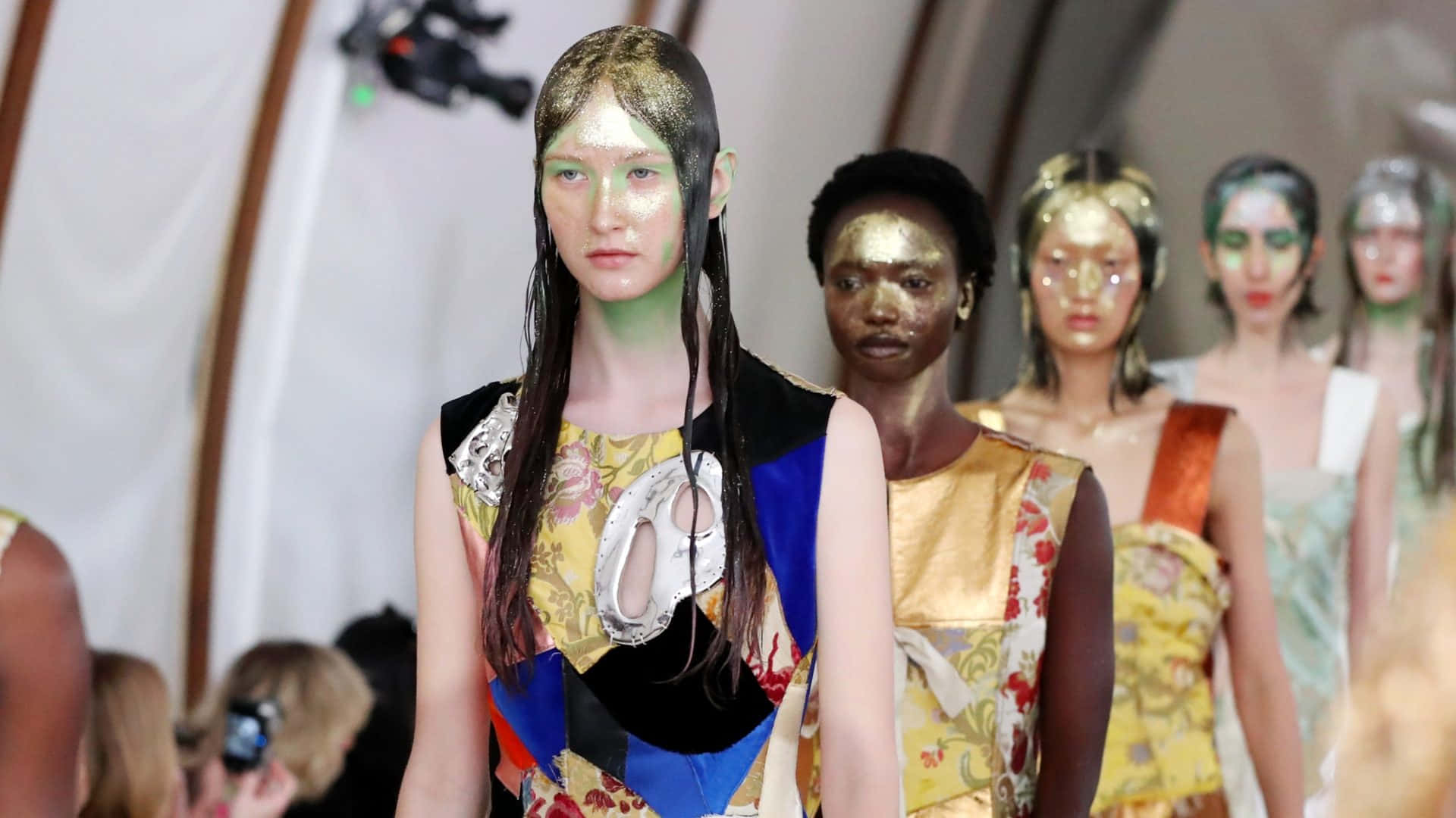 Gold Glitter 桌布 壁紙 — 模特兒臉上塗有金屬金色和綠色顏料，在明亮燈光下穿著大膽圖案服裝走秀。