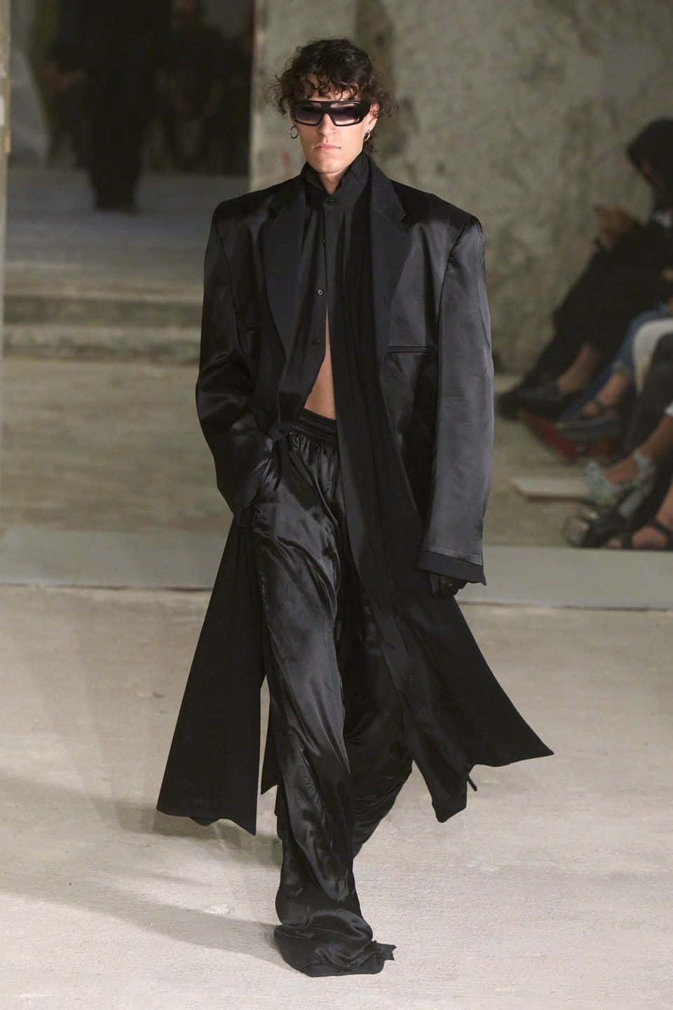 Vetements 桌布 壁紙 — 模特兒自信地走在伸展台上，身穿時尚的全黑緞面套裝，搭配 oversized 外套和太陽眼鏡。