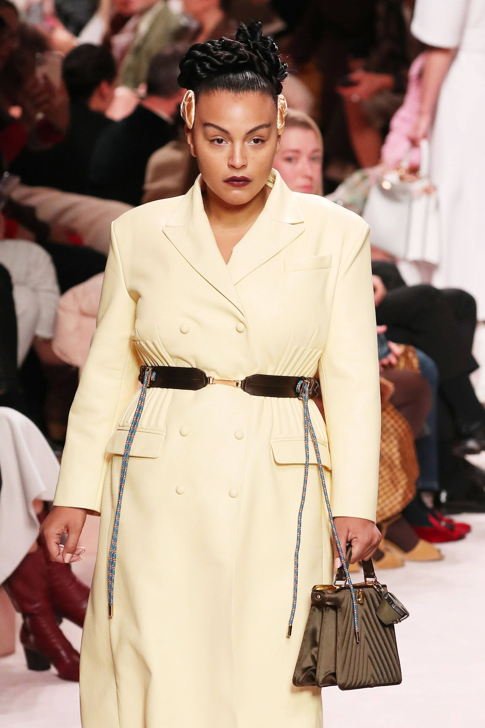 Fendi 桌布 壁紙 — 模特兒自信地走在伸展台上，身穿淡黃色雙排扣大衣，配有寬腰帶，並手持結構感十足的綠色手提包。