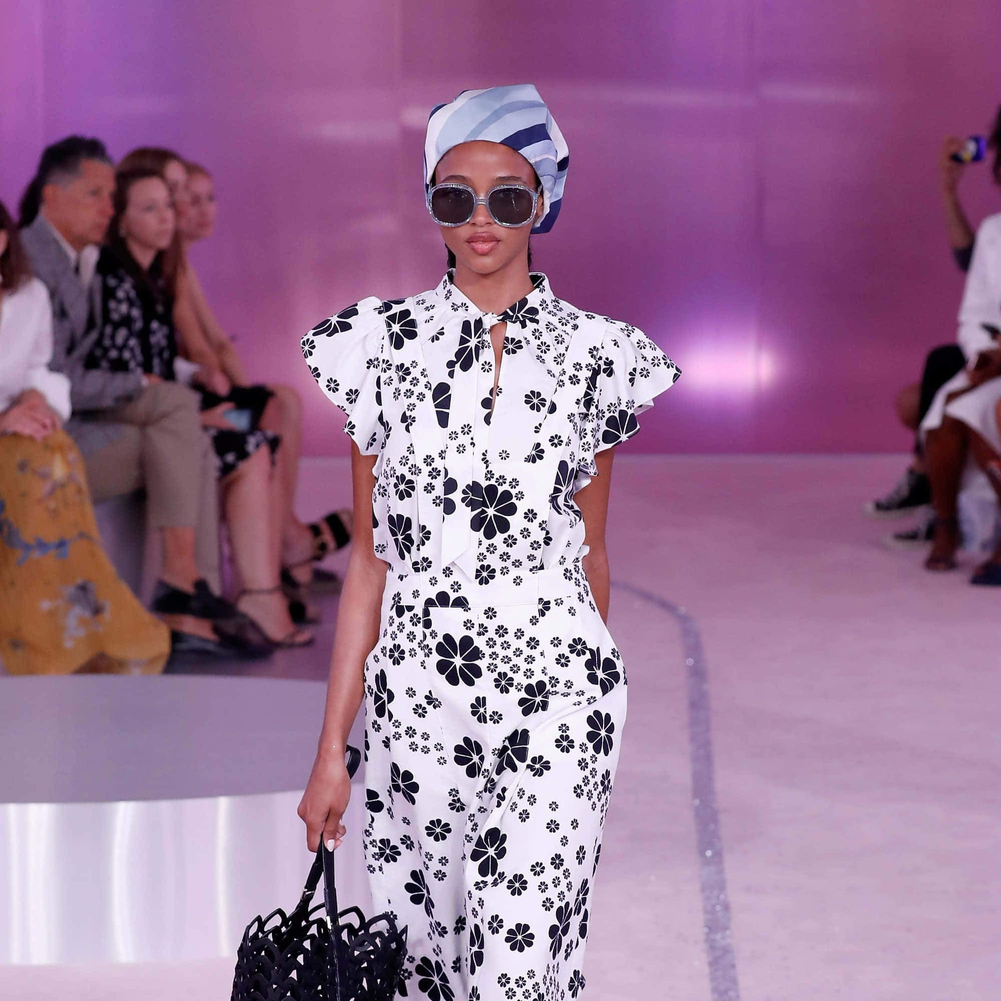 kate spade 桌布 壁紙 — 模特兒自信地走在伸展台上，身穿白色花卉連身褲和條紋頭巾，在紫色燈光下。
