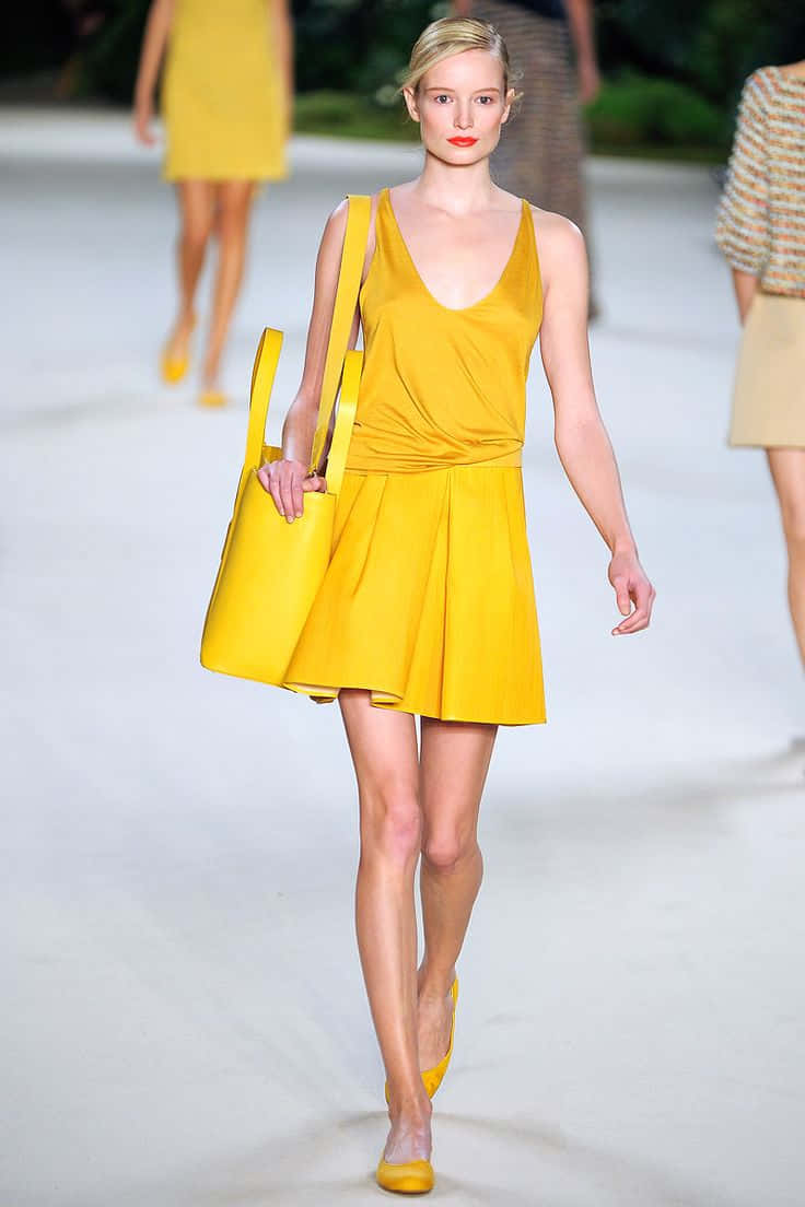 Yellow Fashion 桌布 壁紙 — 模特兒自信地走在伸展台上，身穿鮮黃色無袖連衣裙和相配的手袋，在明亮的攝影棚燈光下。