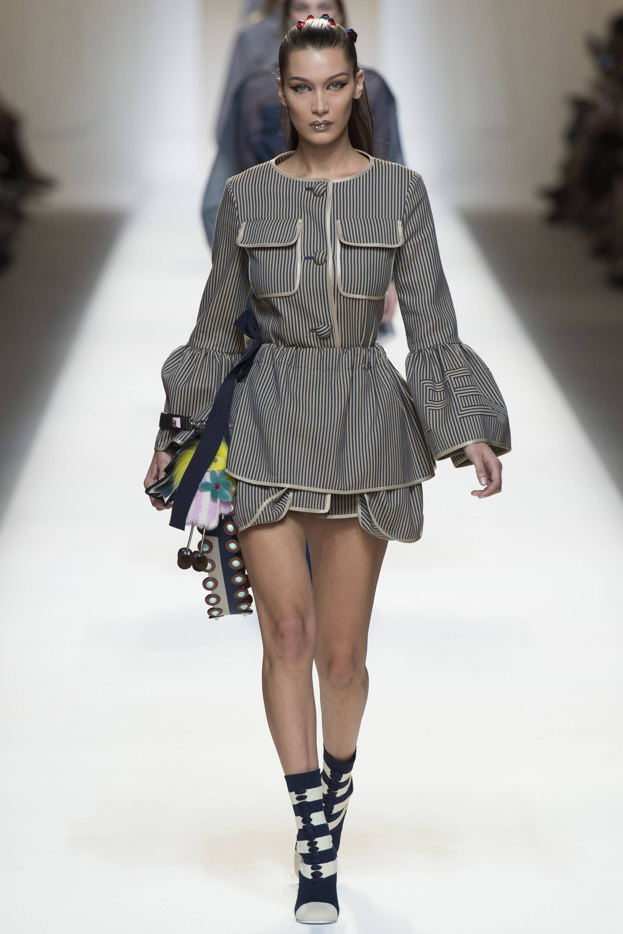 Fendi 桌布 壁紙 — 模特兒自信地走在明亮的白色伸展台上，身穿大膽的條紋荷葉邊上衣，搭配喇叭袖和圖案靴子。