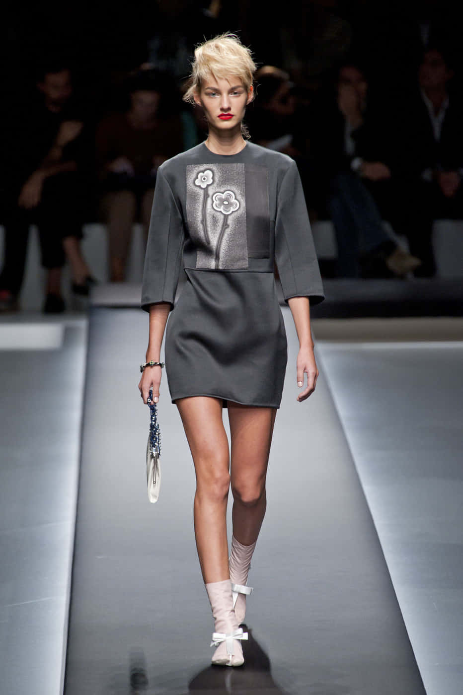 Prada 桌布 壁紙 — 模特兒自信地走在時尚天橋上，身穿灰色刺繡花卉連衣裙，手拿手拿包，在明亮的舞台燈光下。