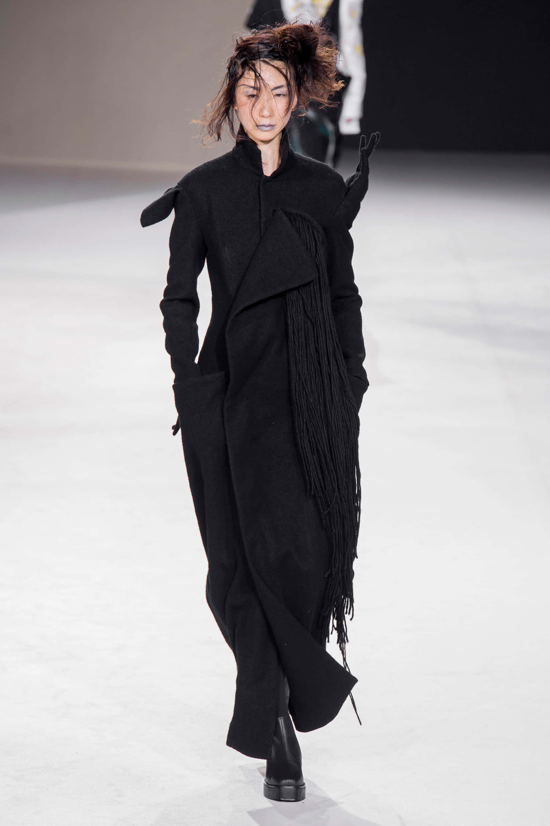 Runway 桌布 壁紙 — 模特兒自信地走在極簡主義的伸展台上，身穿帶有不對稱流蘇和雕塑感肩部的戲劇性黑色大衣。