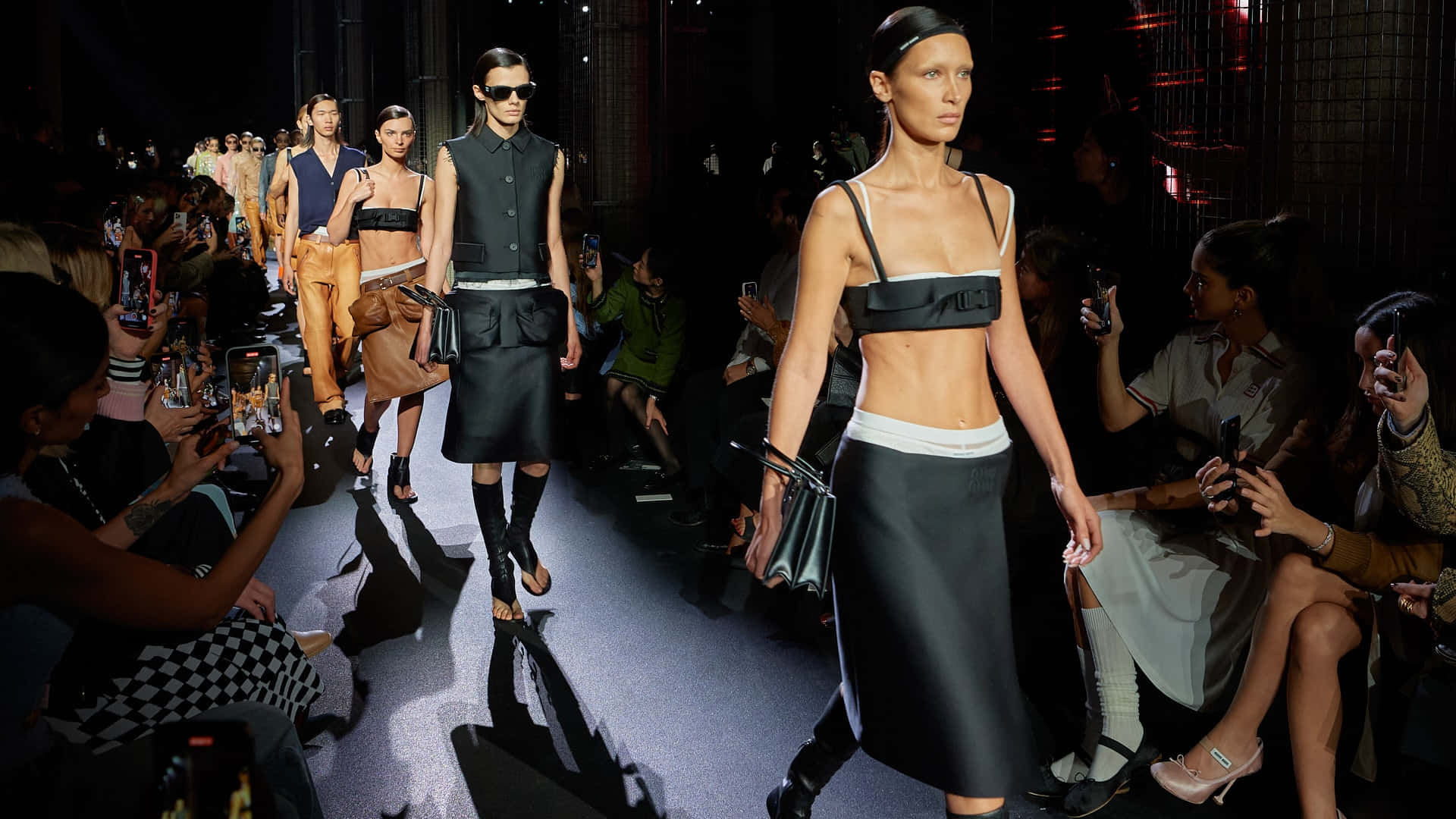 Runway 桌布 壁紙 — 模特兒自信地走在燈光昏暗的時裝伸展台上，身穿前衛黑色服裝，觀眾則用手機捕捉這一刻。