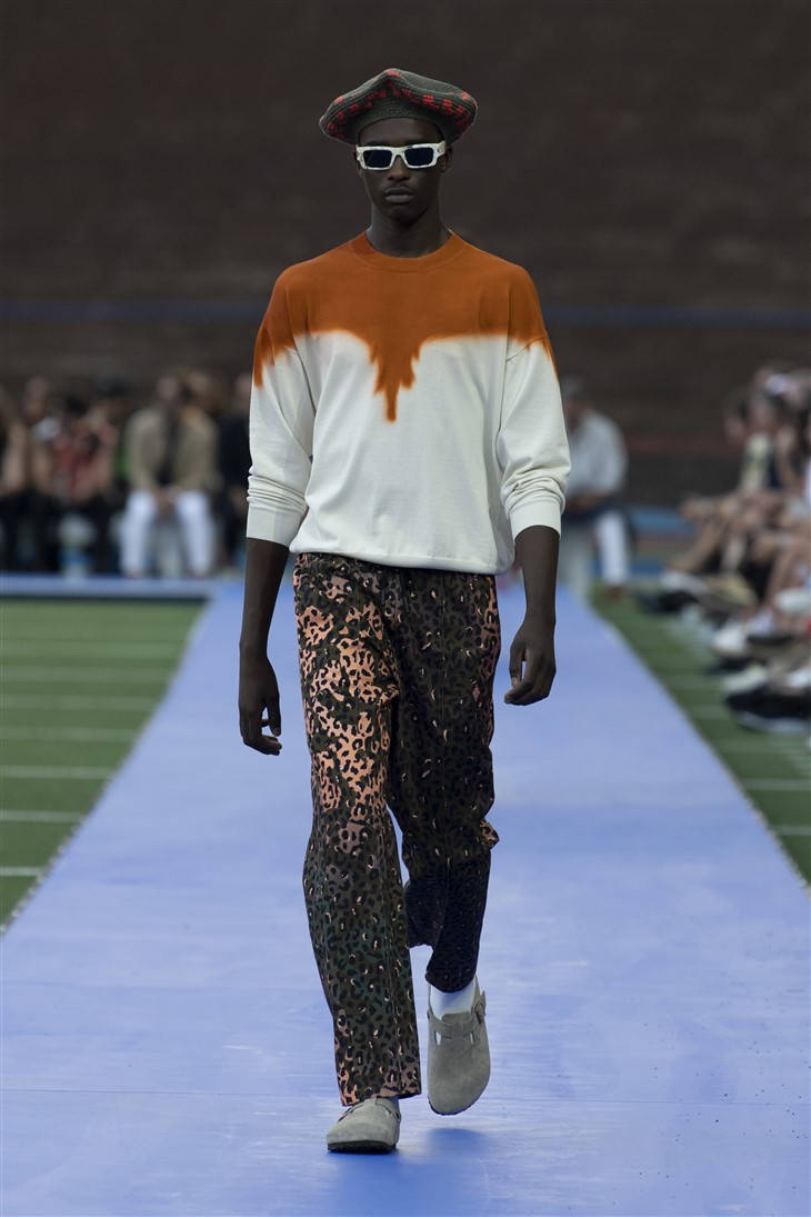 Fashion Model 桌布 壁紙 — 模特兒自信地走在藍色伸展台上，穿著大膽的橙白紮染毛衣、圖案褲子和貝雷帽，在明亮的燈光下。