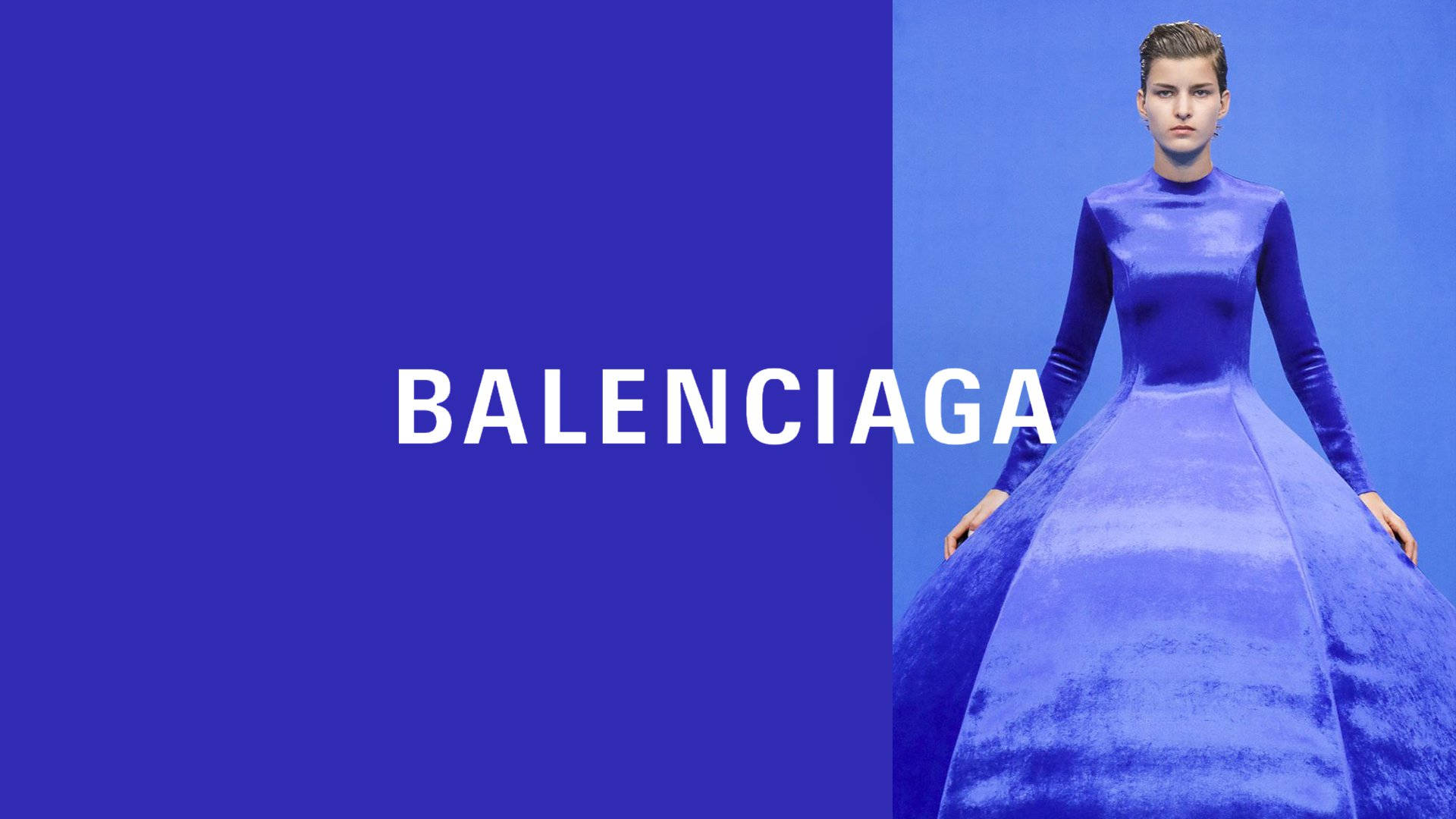 Balenciaga 桌布 壁紙 — 模特兒身穿寬大的皇家藍色長袍，站在帶有 'BALENCIAGA' 文字的藍色分割背景前。