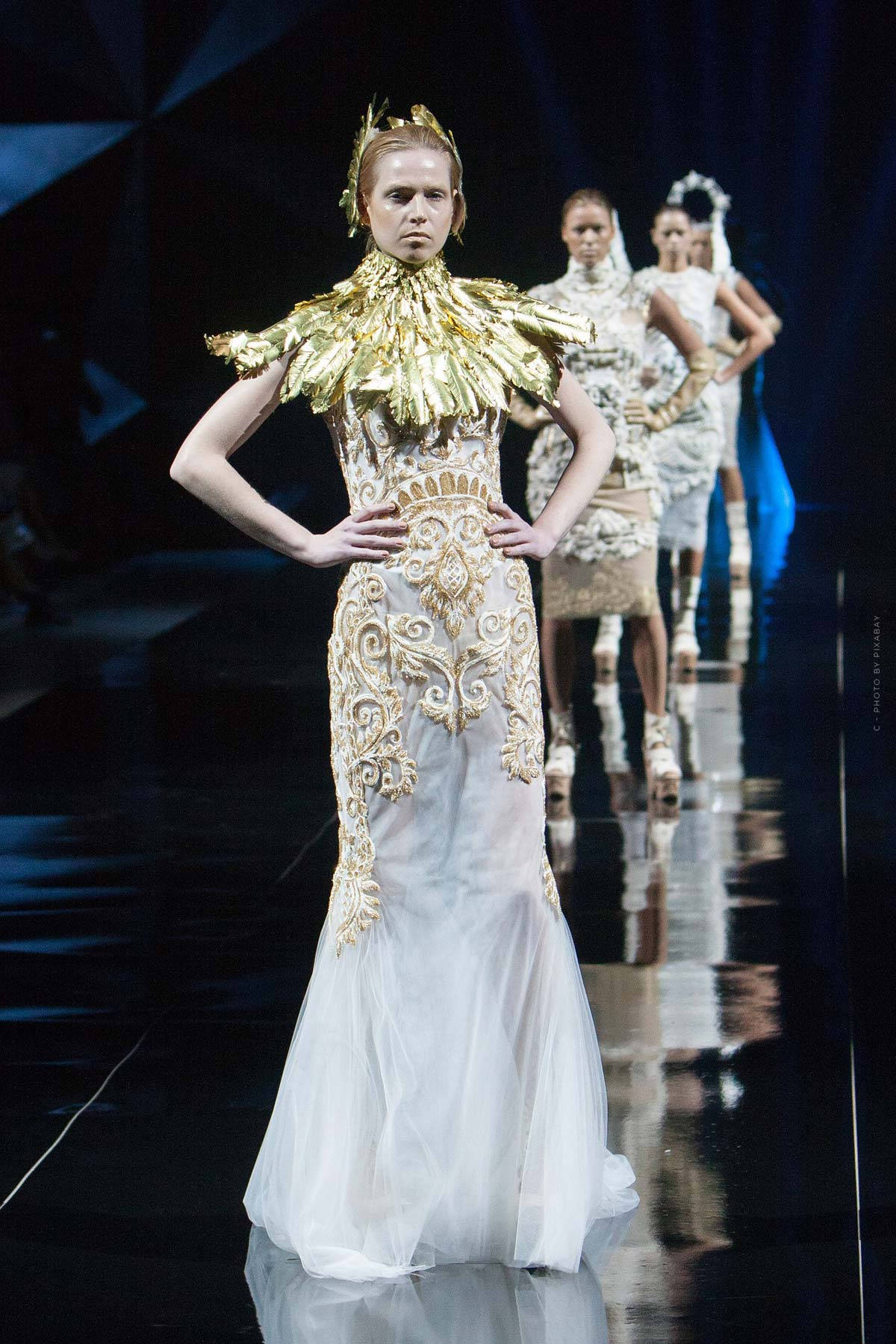 Runway 桌布 壁紙 — 模特兒身穿華麗的金白色禮服，搭配戲劇性的羽毛領，在耀眼的舞台燈光下自信地走在光亮的伸展台上。