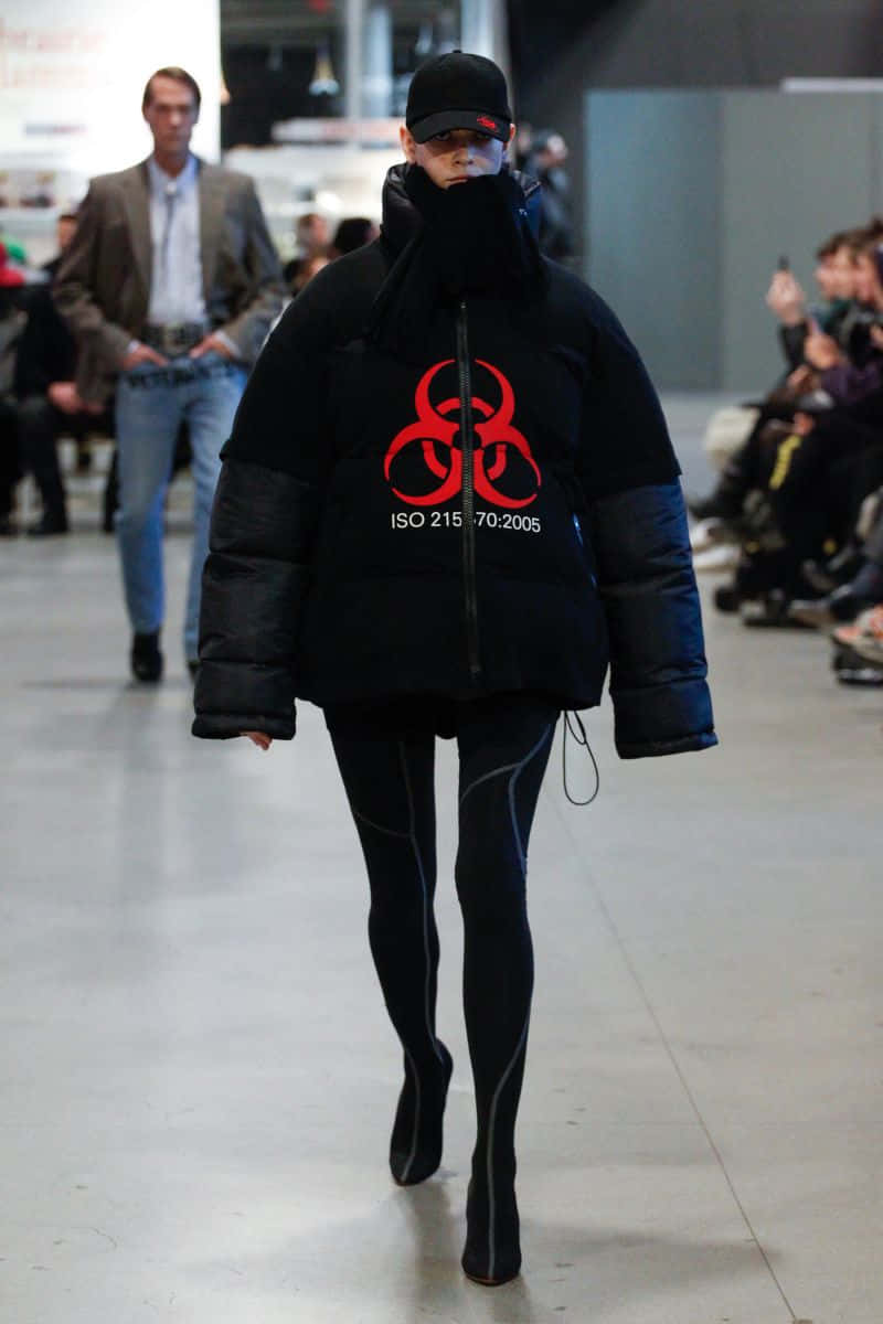 vetements 桌布 壁紙 — 模特兒身穿黑色羽絨外套，上面有紅色生物危害符號和ISO文字，走在伸展台上。