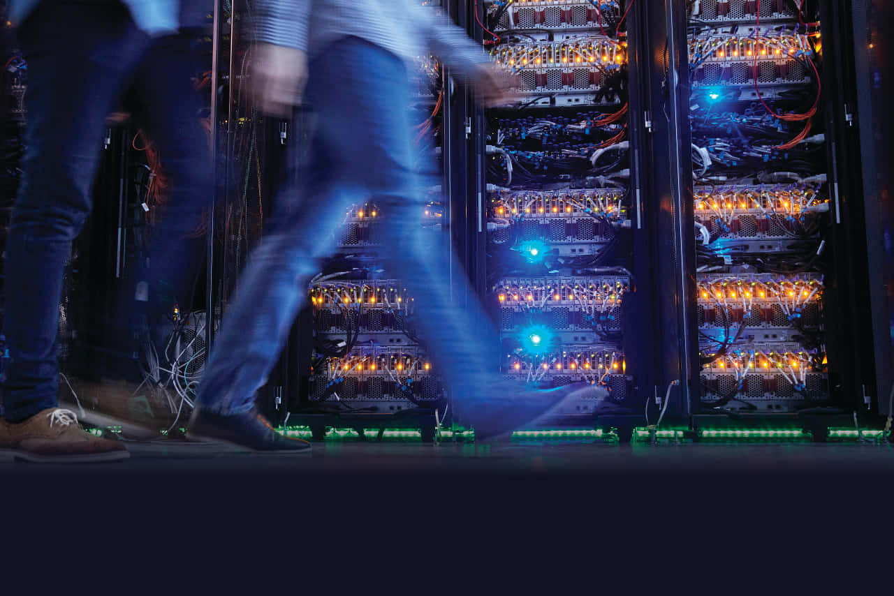 Gaming Servers 桌布 壁紙 — 模糊的技術人員走過昏暗資料中心中發光的伺服器機架，帶有橙色和藍色燈光。