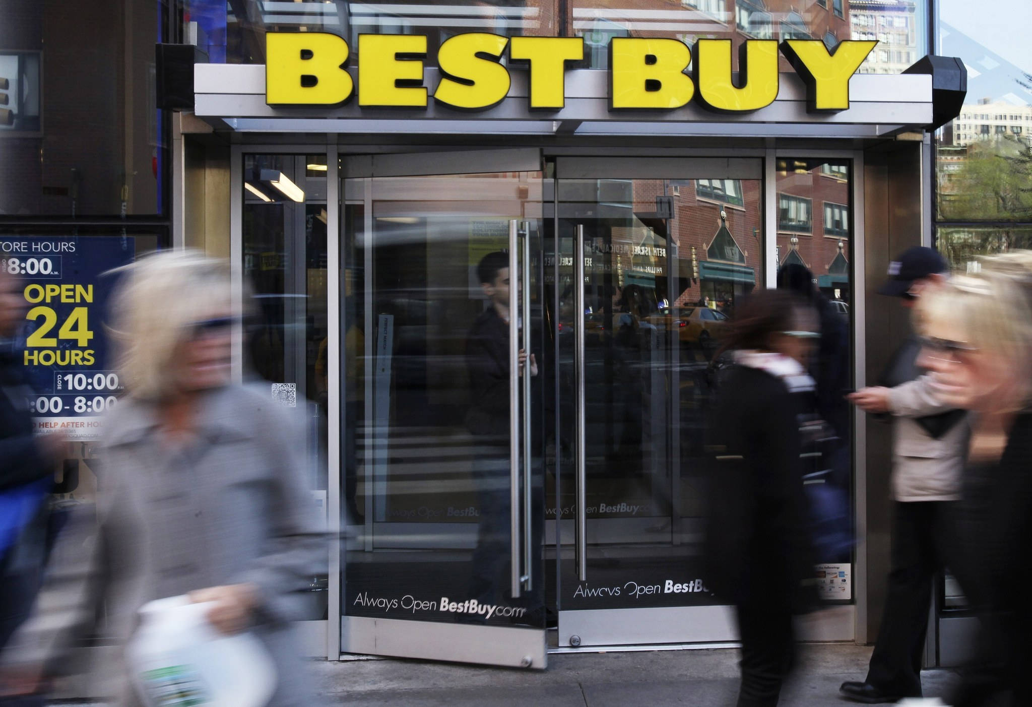 buy 桌布 壁紙 — 模糊的行人匆匆經過Best Buy商店入口，入口有醒目的黃色標誌和玻璃門，反射出城市建築。