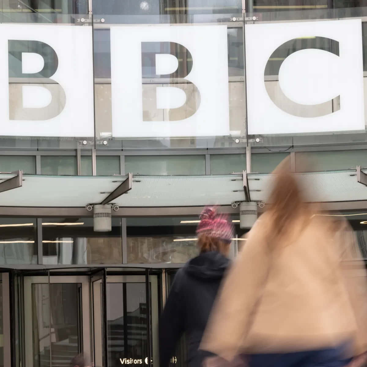 women 桌布 壁紙 — 模糊的行人走過標誌性的BBC大樓外牆，大型玻璃字母反射城市環境。