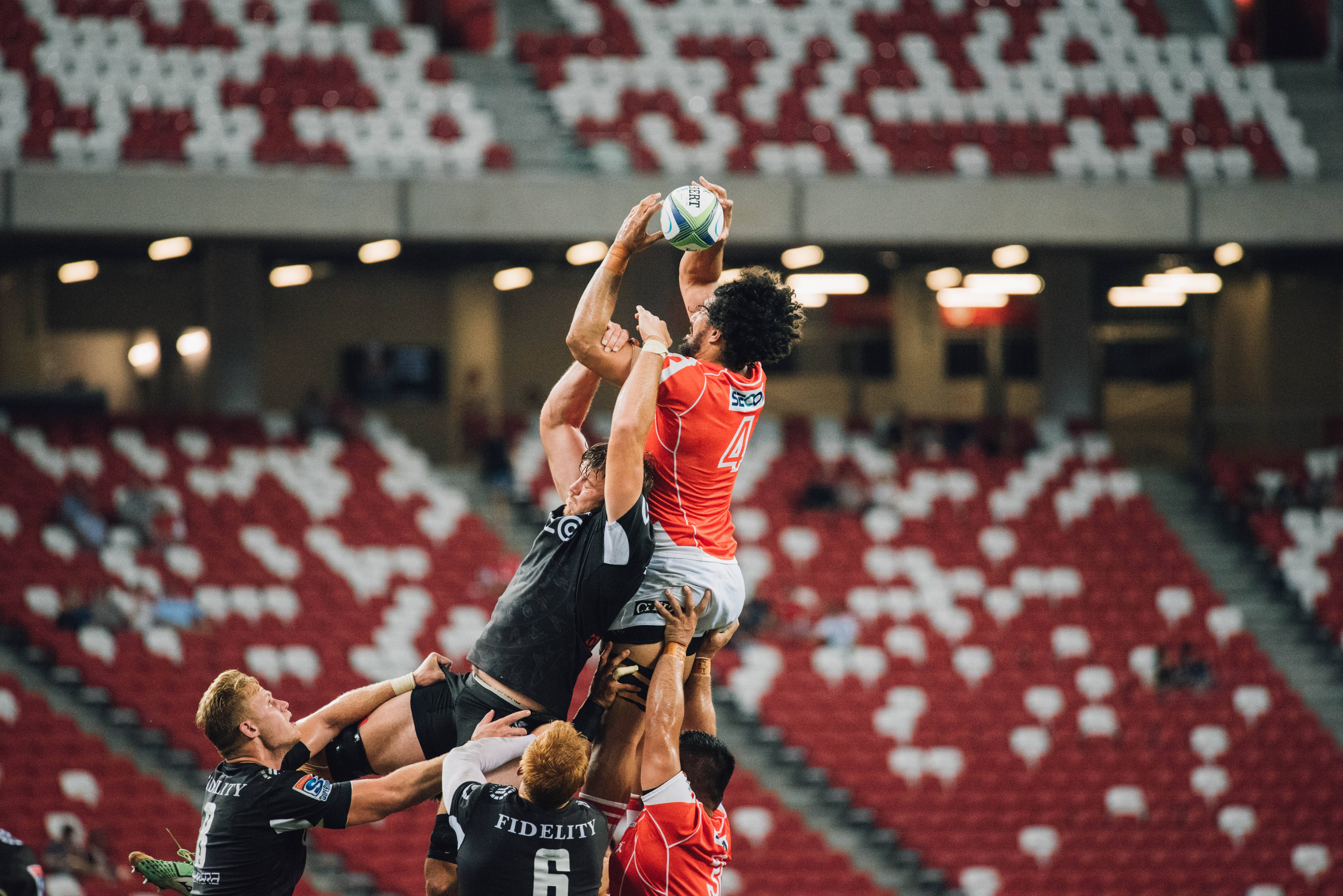 Handball 桌布 壁紙 — 橄欖球球員在球場燈光下跳起爭球，背景是空蕩蕩的紅色座位。