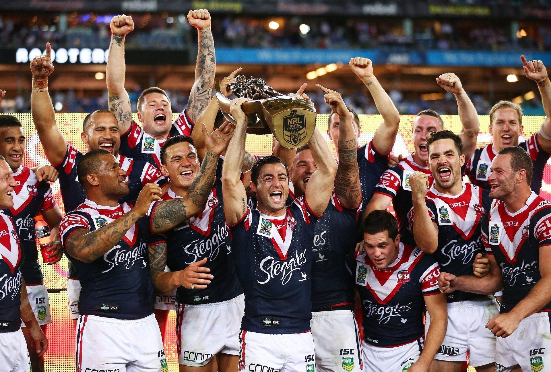 Nrl 桌布 壁紙 — 橄欖球聯賽球員身穿藍、紅、白色球衣，在體育場內瘋狂慶祝，共同舉起一個巨大的金色獎盃。
