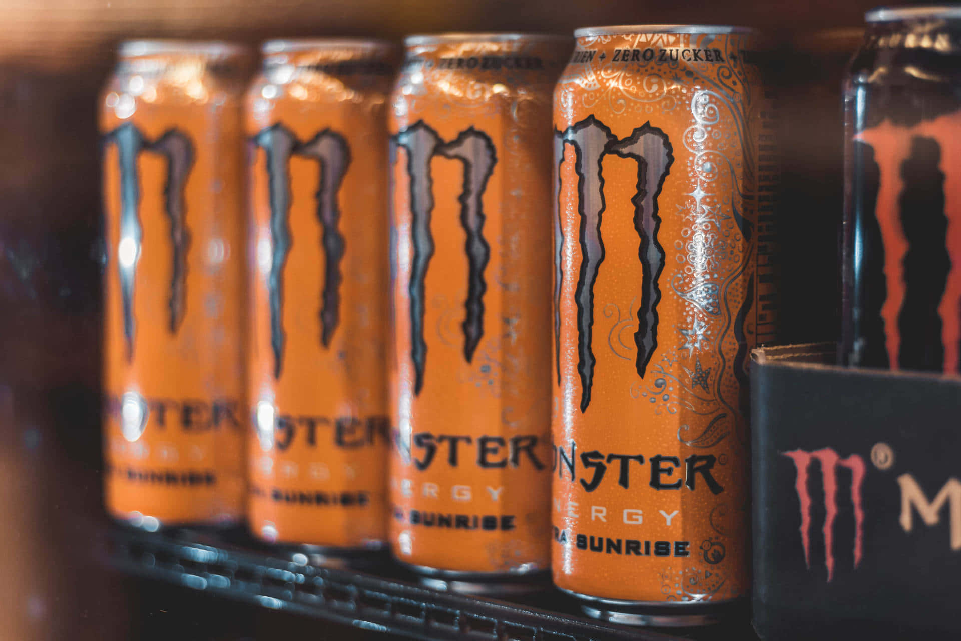 Monster Energy 桌布 壁紙 — 橙色Monster Energy罐頭在貨架上排列的特寫，背景模糊。