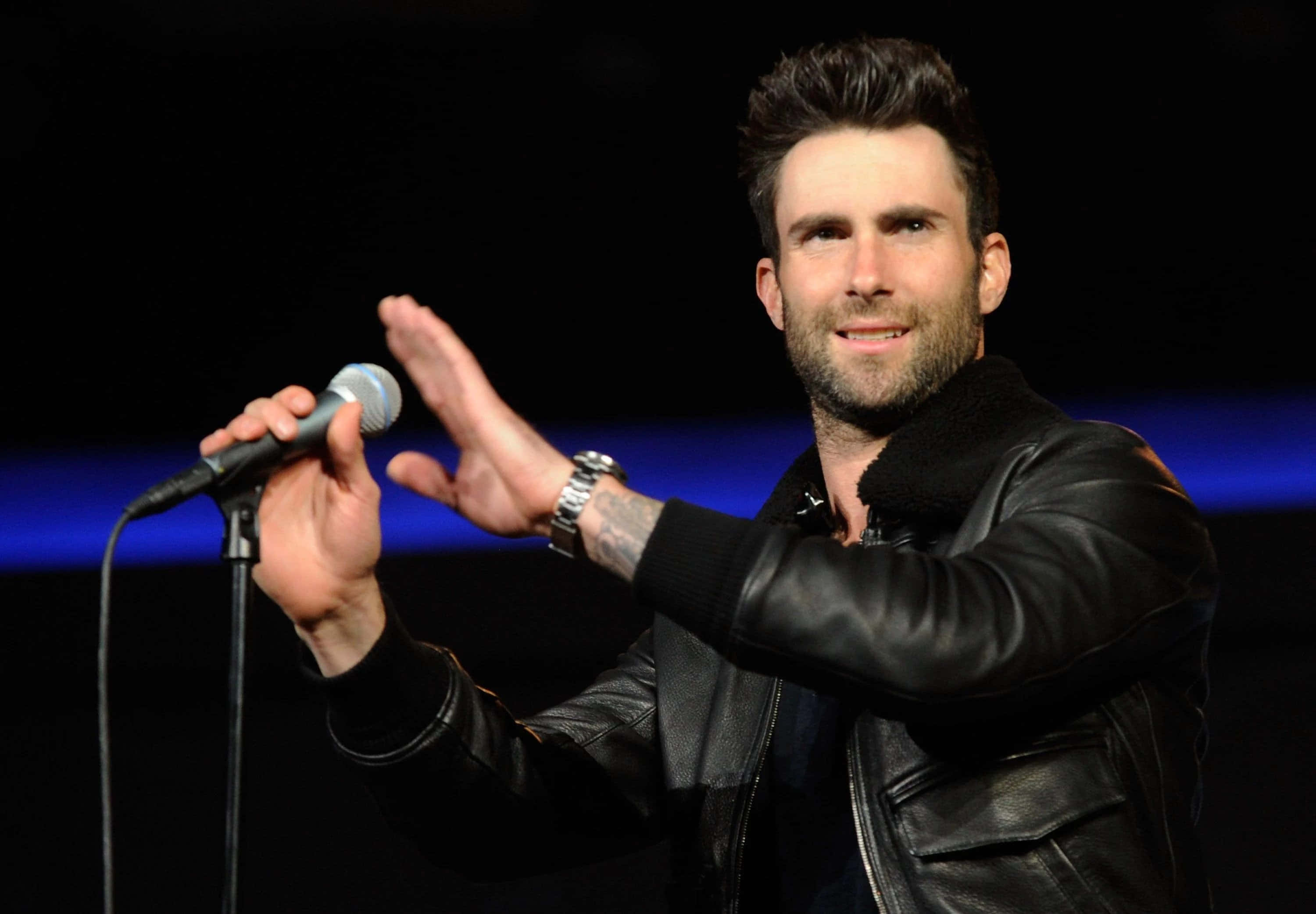 Adam Levine 桌布 壁紙 — 歌手亞當·李維身穿黑色皮夾克在舞台上表演，手持麥克風，溫暖的聚光燈突顯他富有表情的臉龐。