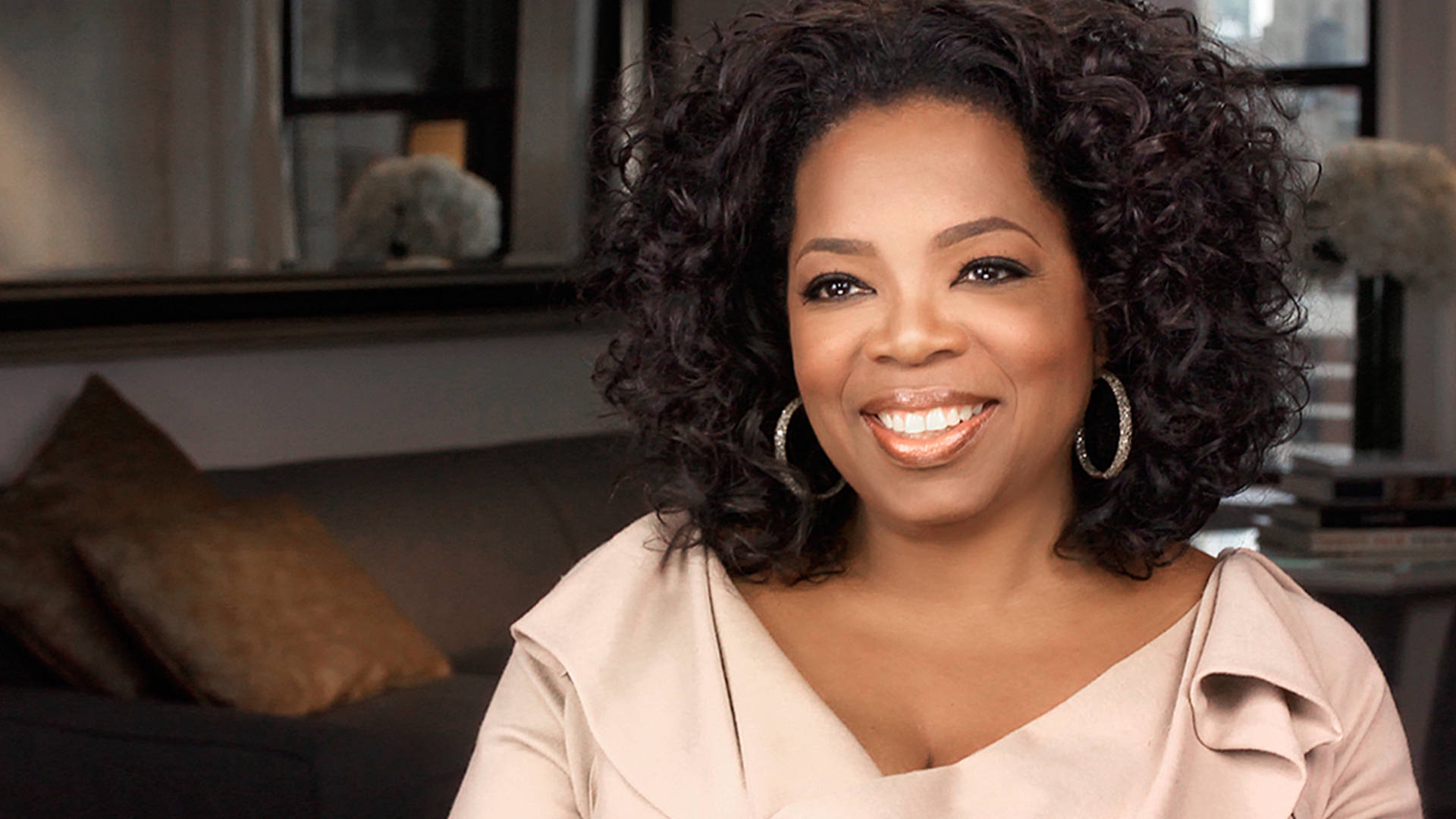 Oprah 桌布 壁紙 — 歐普拉·溫芙瑞在柔和光線的室內環境中溫暖微笑，背景有沙發和窗戶。
