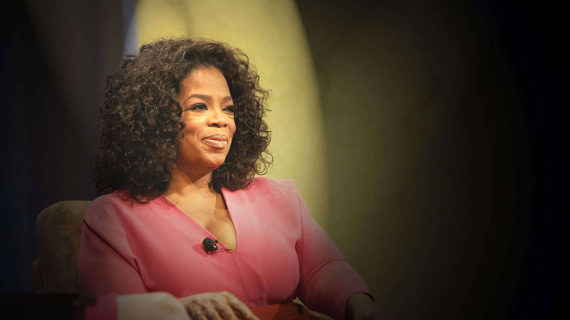 Oprah 桌布 壁紙 — 歐普拉·溫芙瑞在柔和舞台燈光下穿著粉紅色襯衫微笑，背景模糊。