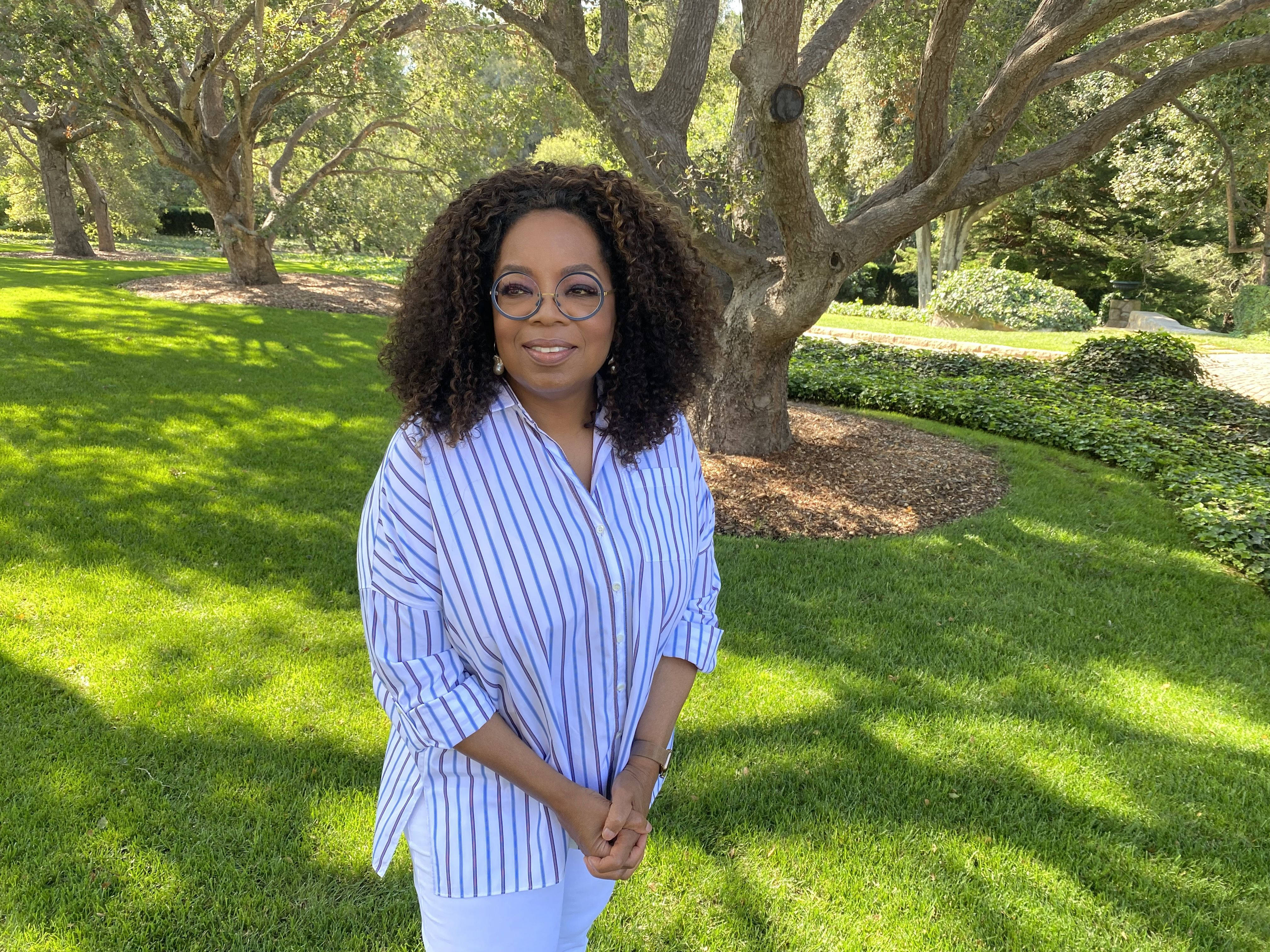 Oprah 桌布 壁紙 — 歐普拉·溫芙蕾（Oprah Winfrey）身穿條紋襯衫，站在枝葉茂密的樹下，在陽光明媚的綠色草坪上溫暖地微笑。