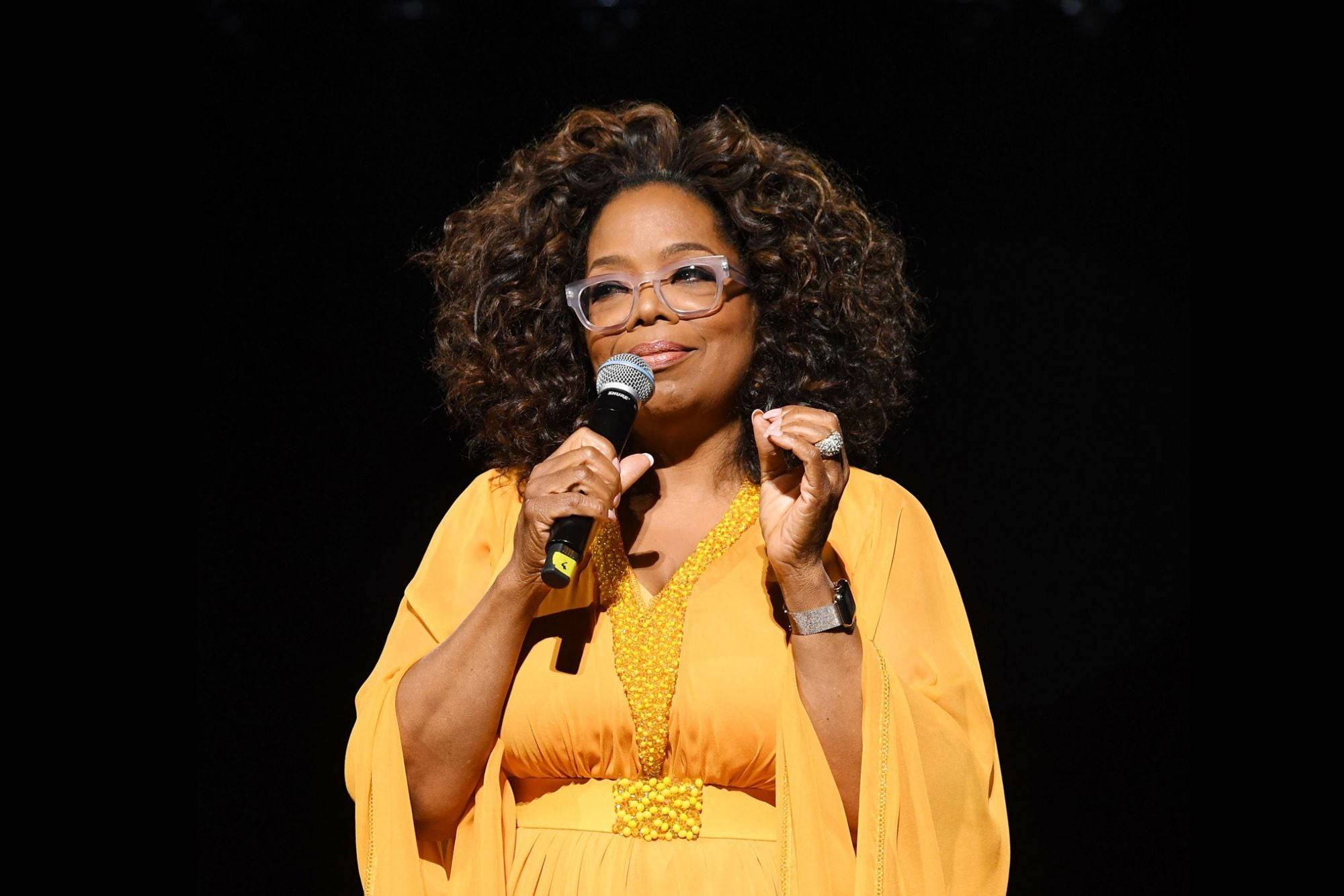 Oprah 桌布 壁紙 — 歐普拉·溫芙蕾在舞台燈光下穿著鮮黃色連衣裙對著麥克風說話。