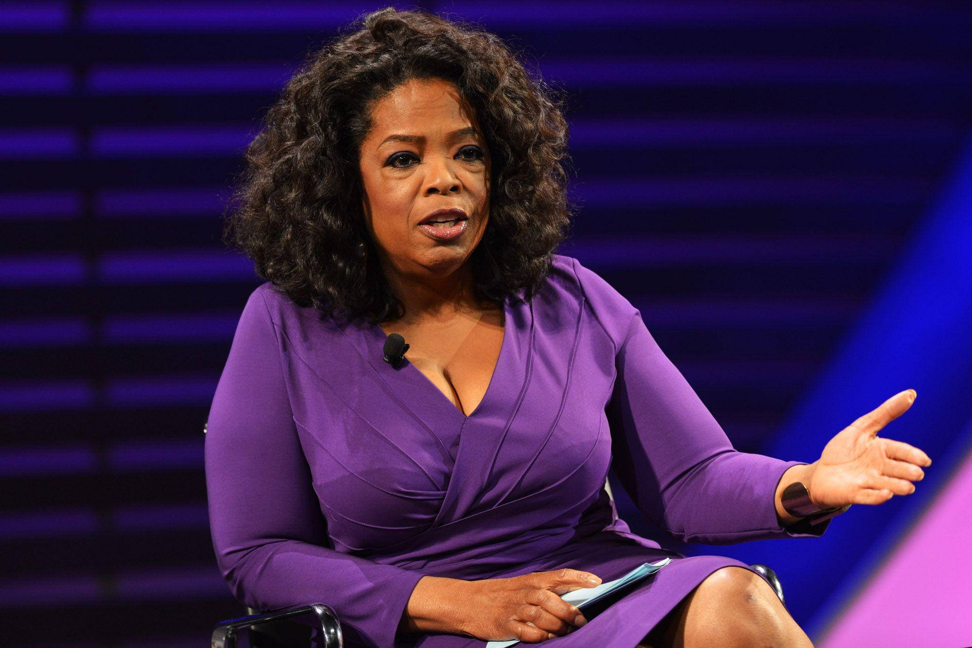 Oprah 桌布 壁紙 — 歐普拉·溫芙蕾穿著鮮豔的紫色連衣裙，在動態的藍色和紫色舞台背景下熱情地演講。