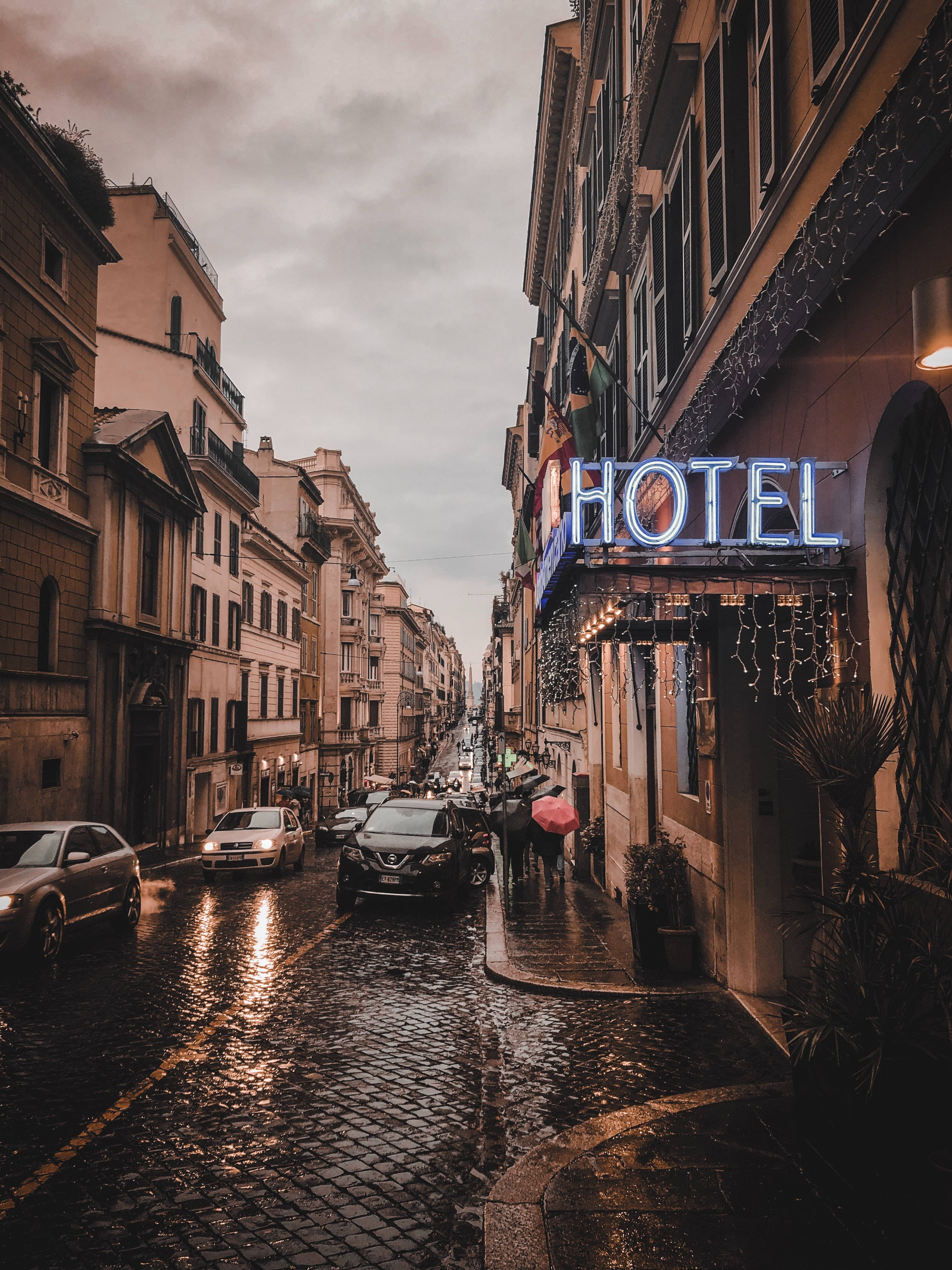 Hotel 桌布 壁紙 — 歐洲城市一條雨中的鵝卵石街道，帶有發光的藍色飯店招牌和撐傘的行人。