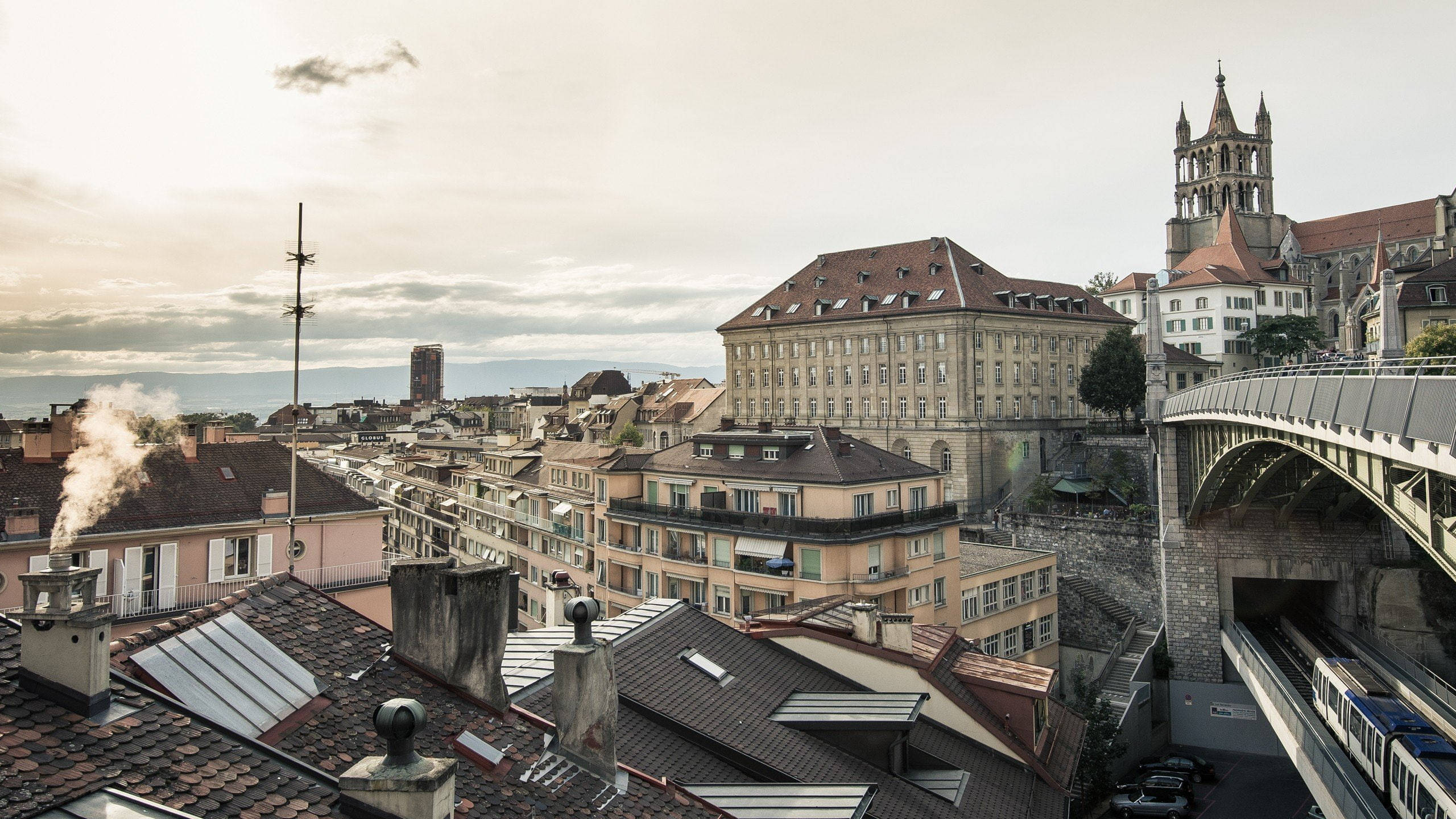 Switzerland 桌布 壁紙 — 歐洲城市景觀的全景視圖，包括歷史建築的屋頂和一座橋樑，橋下有火車在陰天駛過。
