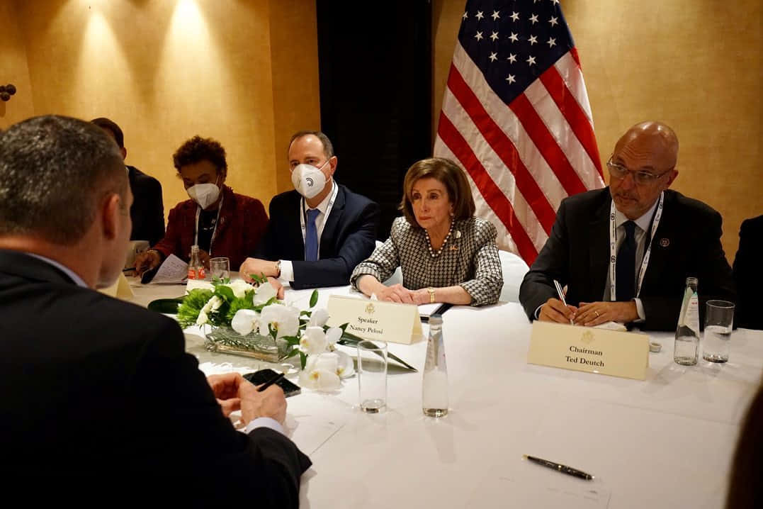 USA Flag 桌布 壁紙 — 正式政治會議，官員圍坐在桌子旁，桌上有名牌，背景有美國國旗。