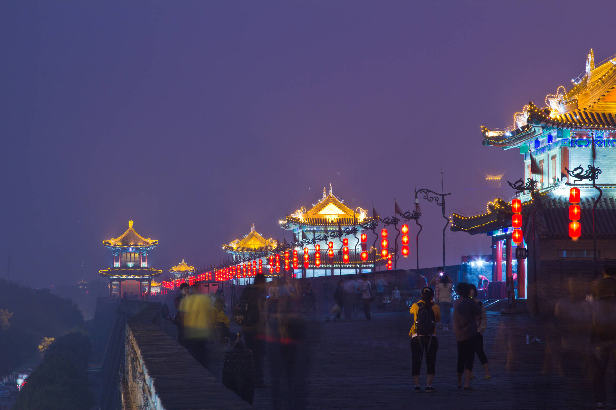 Xian 桌布 壁紙 — 歷史中國城牆的夜景，金色燈籠和閃爍的寶塔屋頂照亮，模糊的行人沿著小路行走。