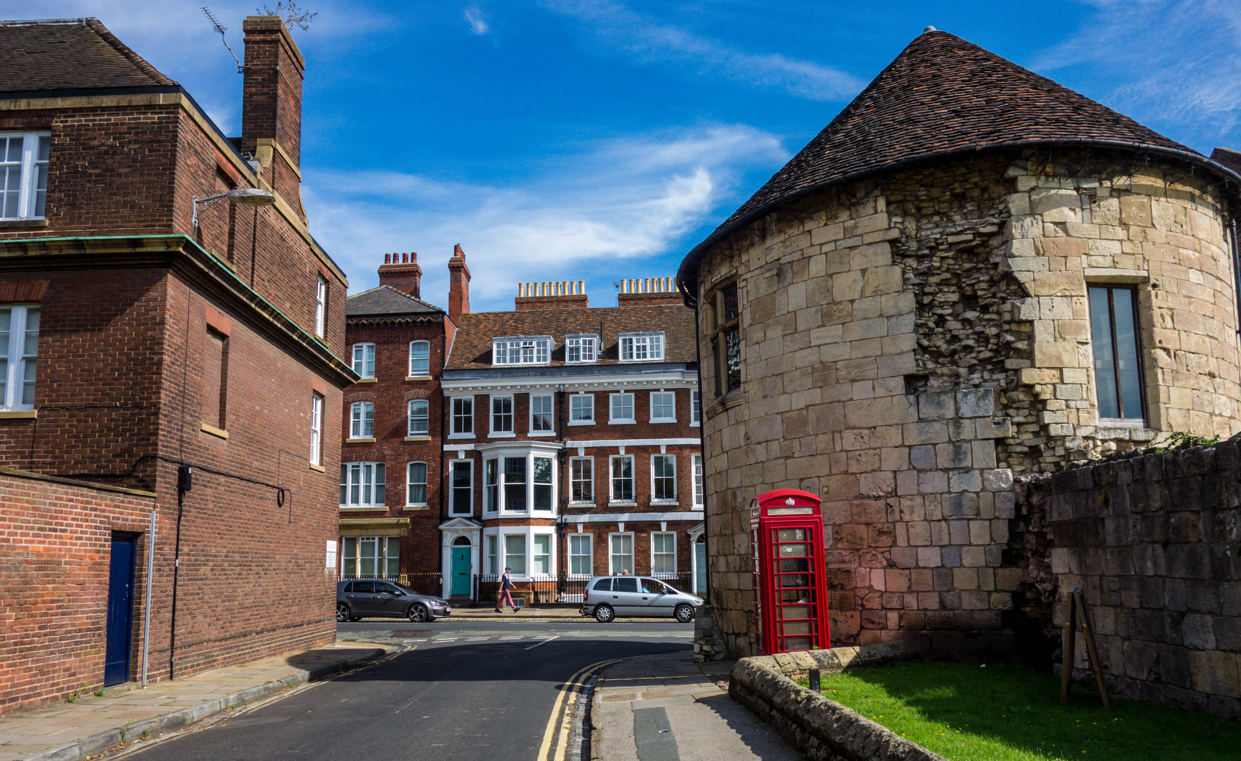 England 桌布 壁紙 — 歷史小鎮的陽光街角，一座紅色電話亭旁邊是風化的石塔和磚建築，背景是清澈的藍天。
