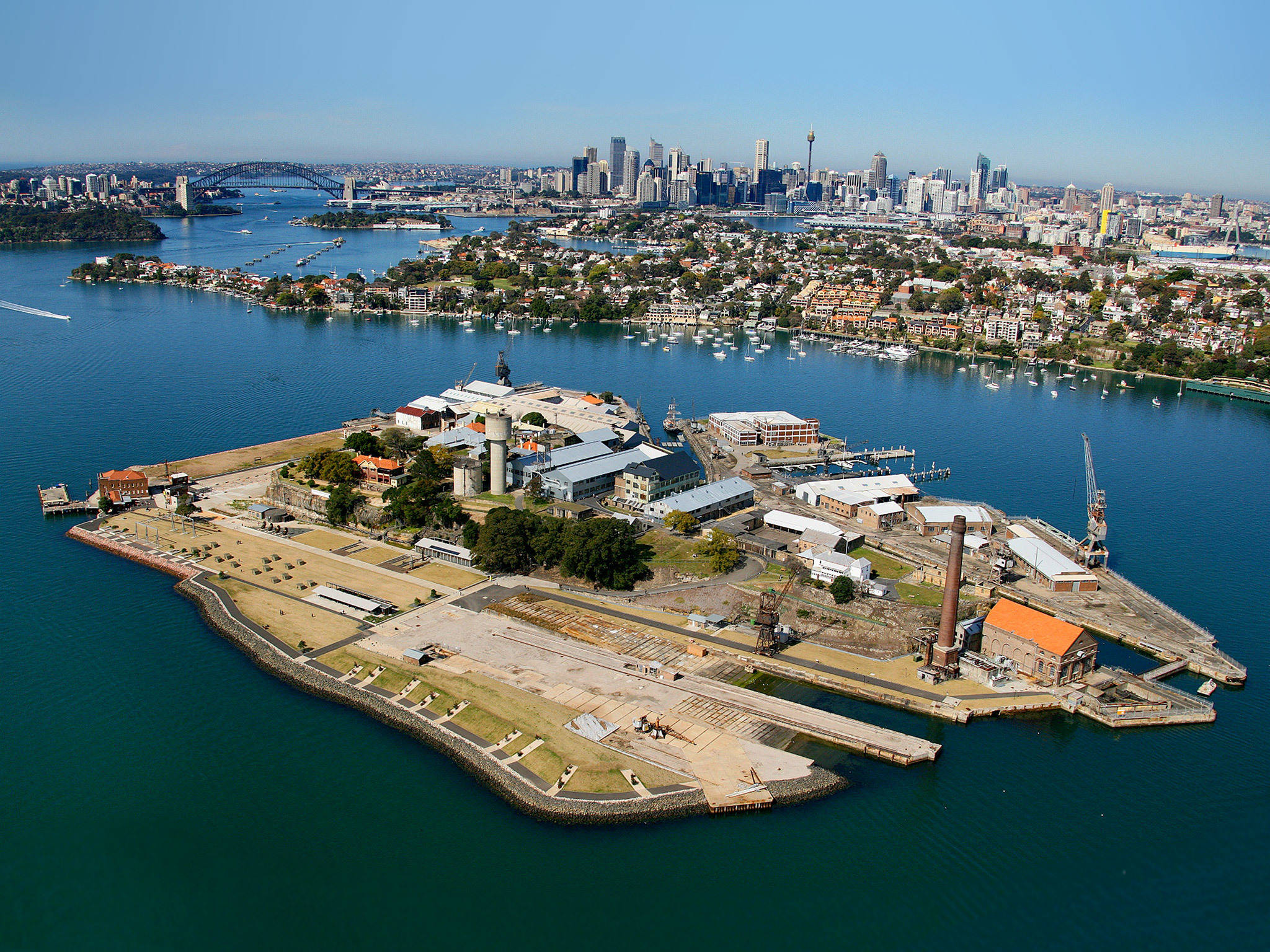 Sydney 桌布 壁紙 — 歷史島嶼堡壘的鳥瞰圖，工業建築被深藍色水域環繞，遠方現代城市天際線在清澈藍天下。