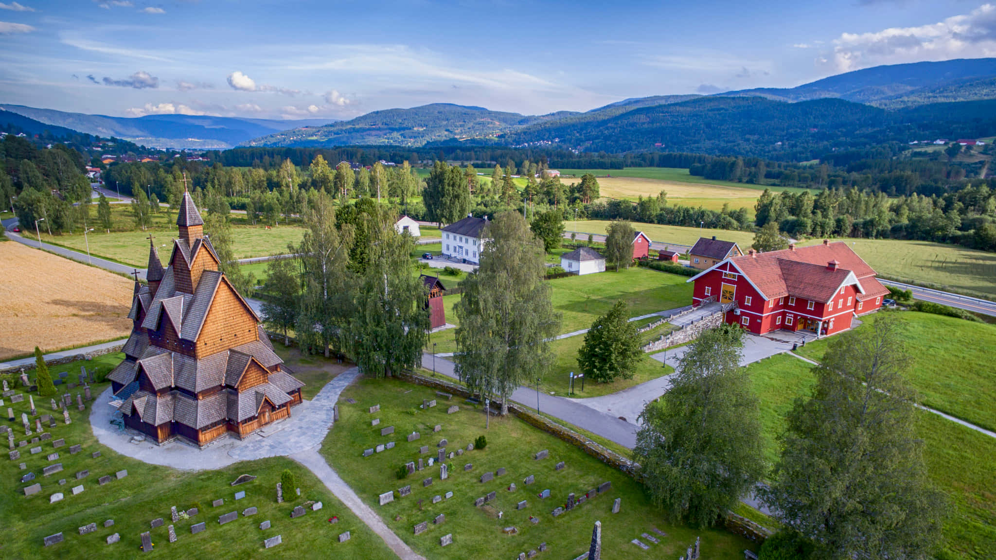 Norway 桌布 壁紙 — 歷史悠久的木造木板教堂的鳥瞰圖，周圍環繞著墓地和紅頂建築，位於鬱鬱蔥蔥的綠色山谷中。