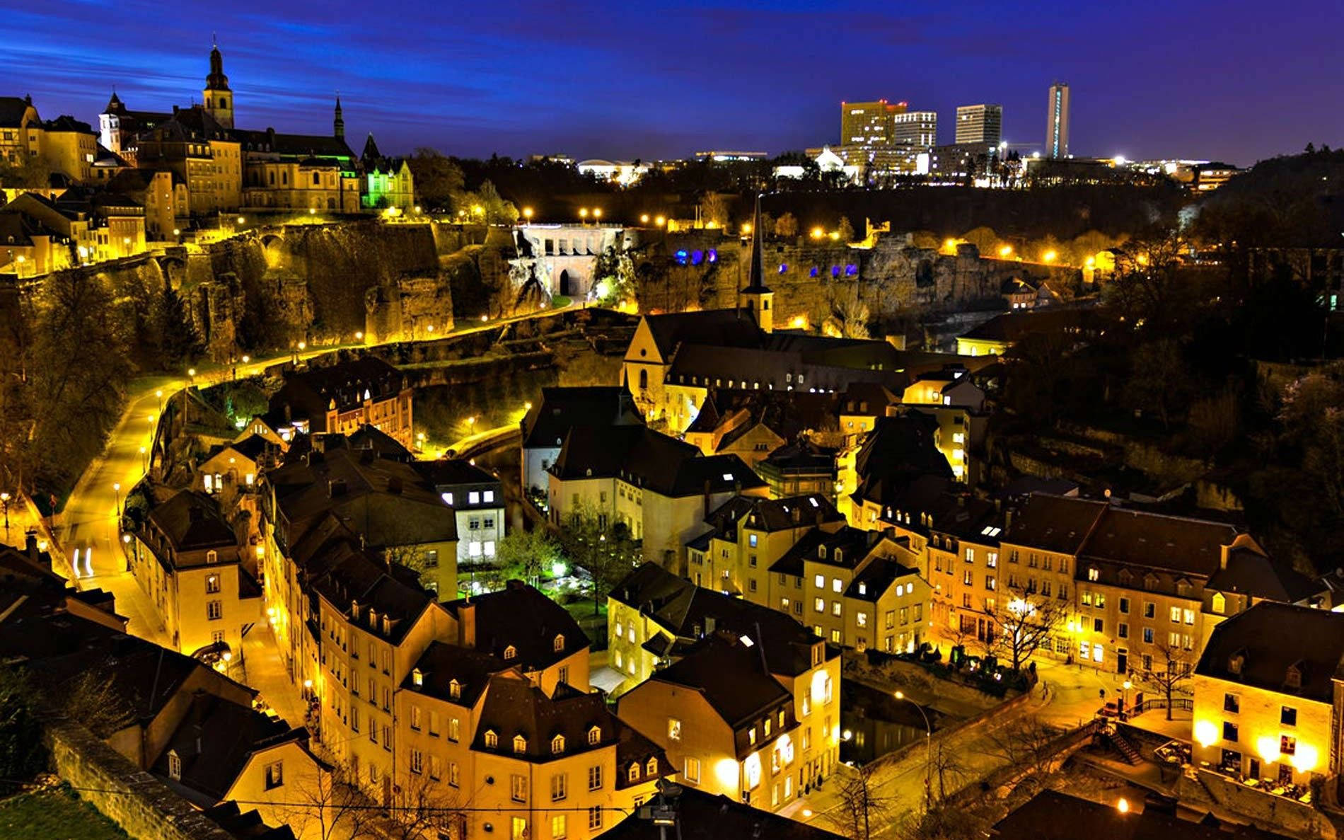 Luxembourg 桌布 壁紙 — 歷史悠久的歐洲城市夜景鳥瞰圖，溫暖的燈光映照著中世紀建築和現代摩天大樓，背景是深藍色的天空。