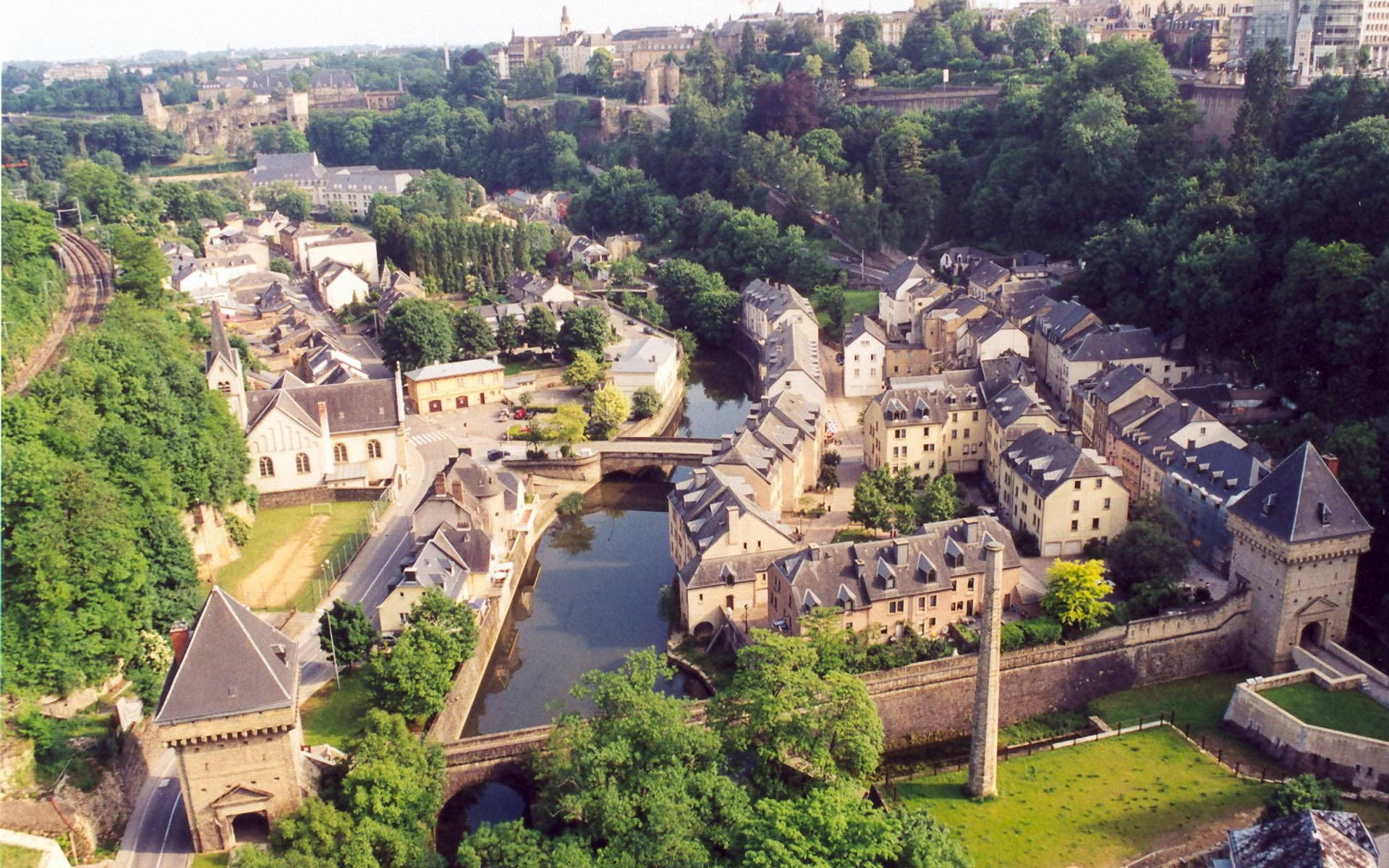 Luxembourg 桌布 壁紙 — 歷史悠久的歐洲小鎮的鳥瞰圖，坐落在蜿蜒的河流旁，周圍環繞著茂密的綠樹和石頭建築。