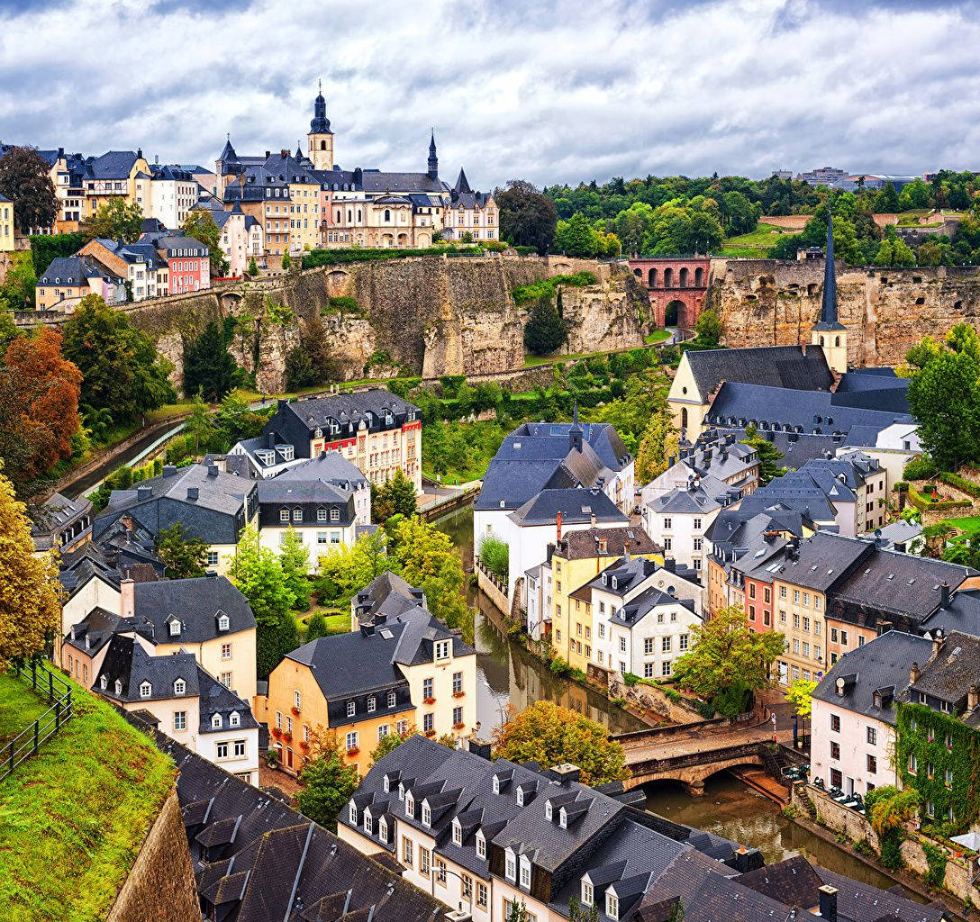 Luxembourg 桌布 壁紙 — 歷史悠久的歐洲小鎮鳥瞰圖，擁有色彩繽紛的建築、石橋和河流，蜿蜒穿過中世紀建築，天空多雲。