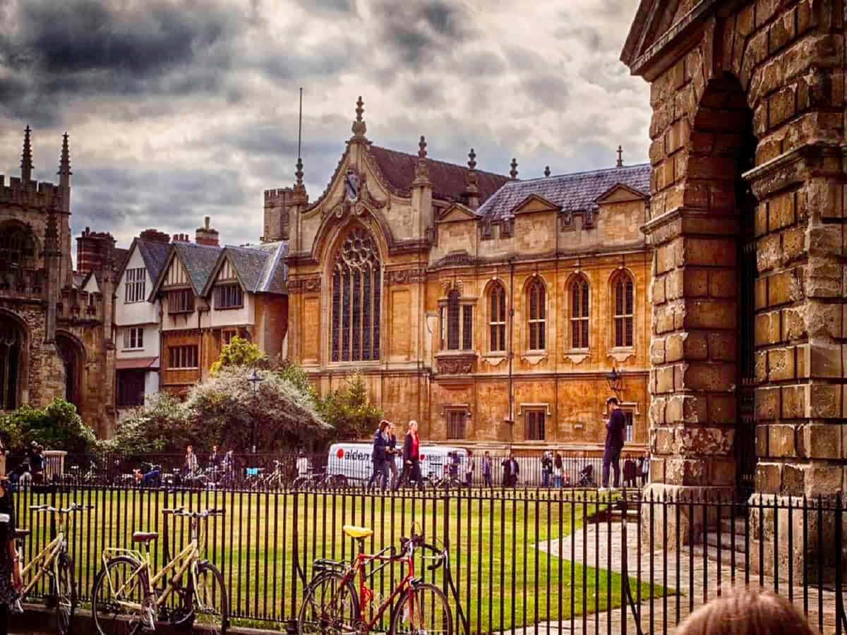 Oxford United Kingdom 桌布 壁紙 — 歷史悠久的石造大學庭院，哥德式建築，腳踏車停放在圍欄旁，人們在陰天行走。