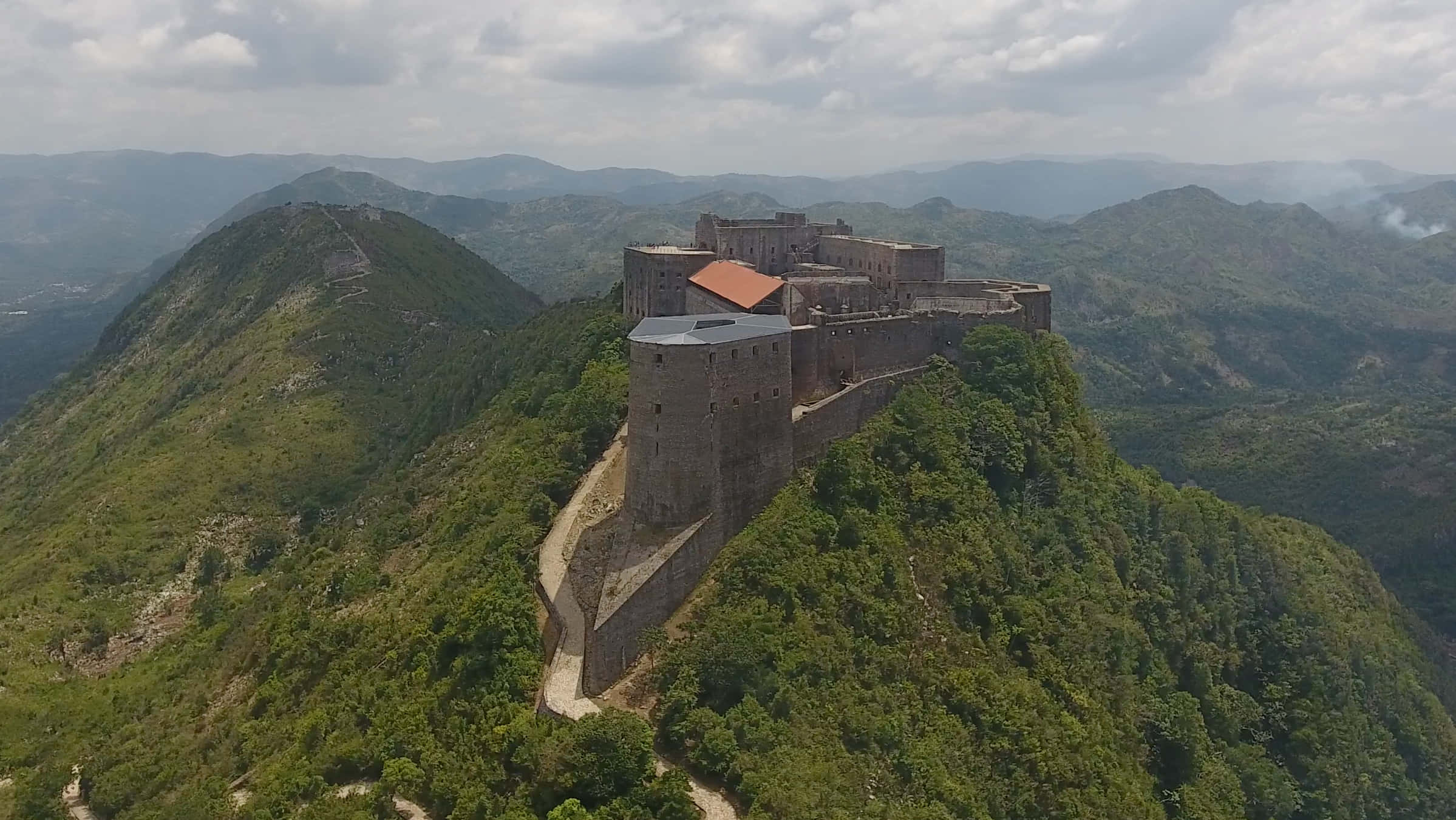 Fortress 桌布 壁紙 — 歷史悠久的石頭堡壘坐落在鬱鬱蔥蔥的山脊上，俯瞰著多雲天空下連綿起伏的丘陵的鳥瞰圖。