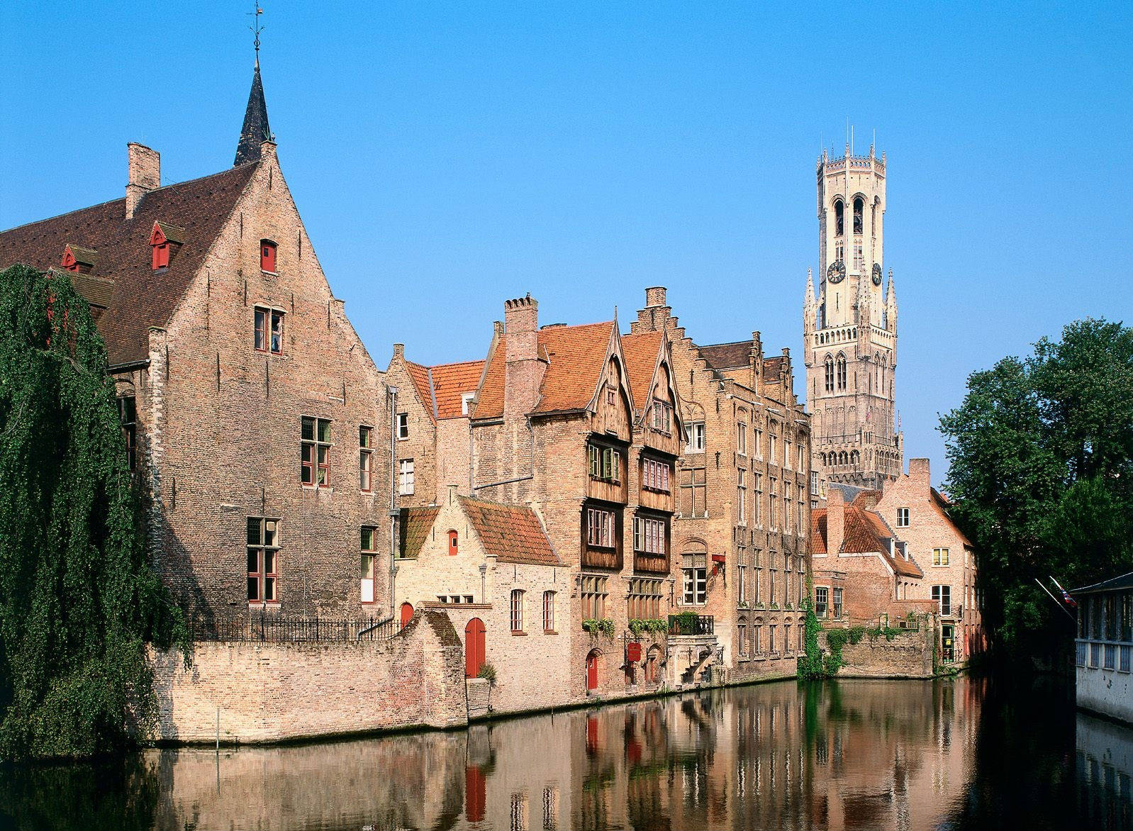 Belgium 桌布 壁紙 — 歷史悠久的磚砌建築，紅色屋頂，沿著平靜的運河排列，在清澈的藍天下，遠處有一座高高的鐘樓。