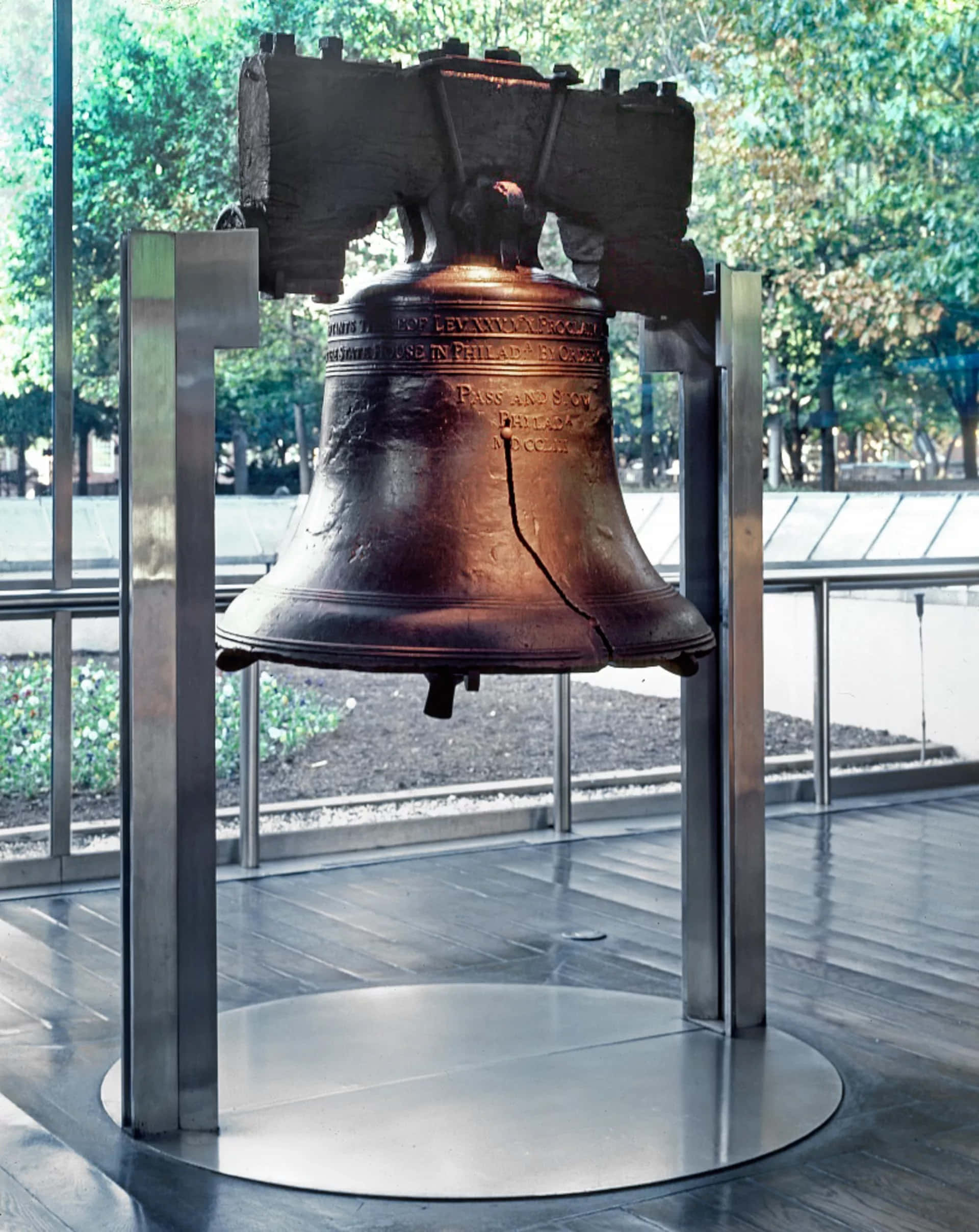 Bell 桌布 壁紙 — 歷史悠久的自由鐘帶有其標誌性裂縫，懸掛在現代玻璃圍欄內，外面可見樹木。
