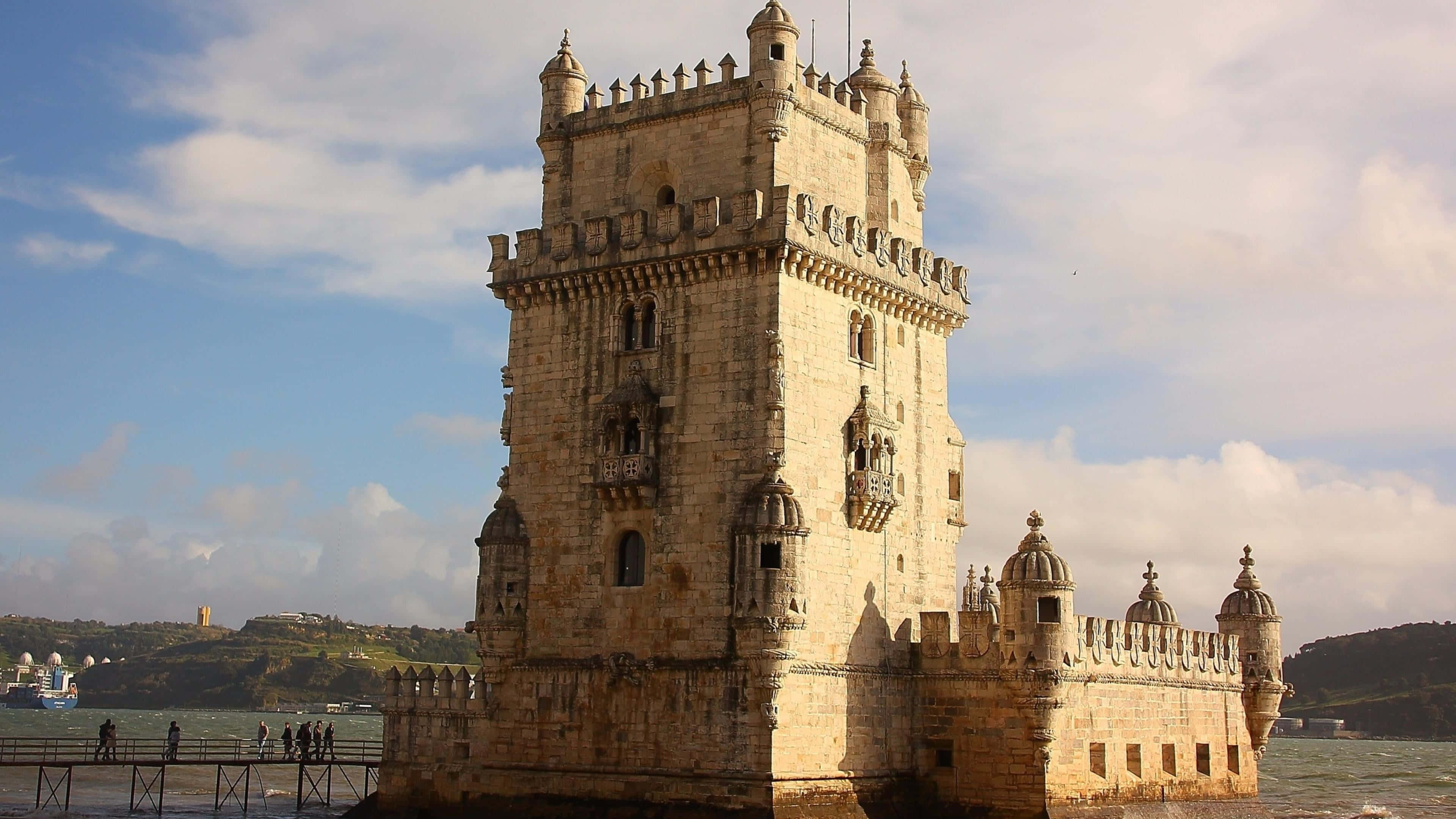 Portugal 桌布 壁紙 — 歷史悠久的貝倫塔在部分多雲的天空下雄偉地矗立在水邊，附近有遊客。