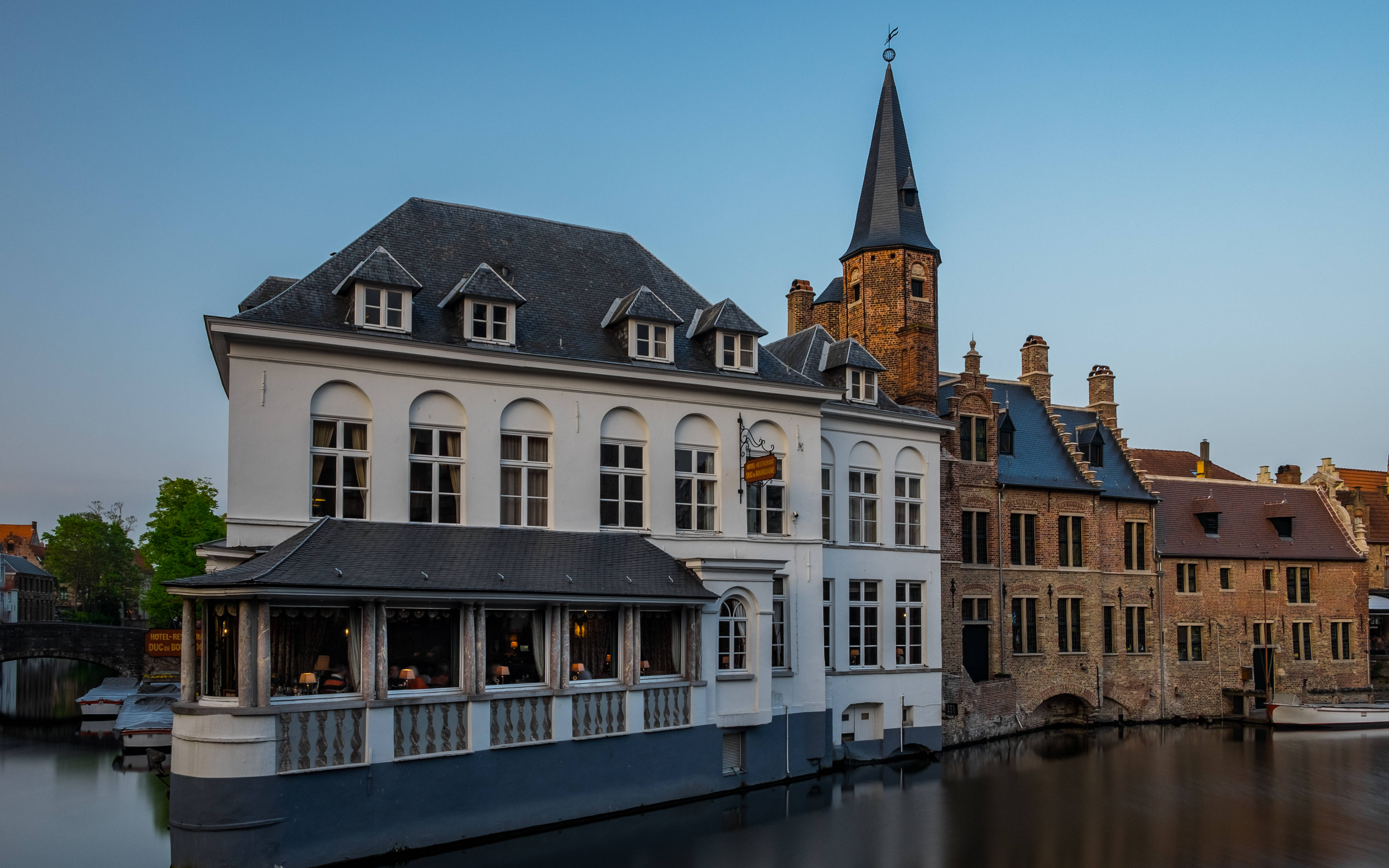 Belgium 桌布 壁紙 — 歷史悠久的運河畔建築，擁有拱形窗戶和石塔，在暮色中倒映在平靜的水面上。