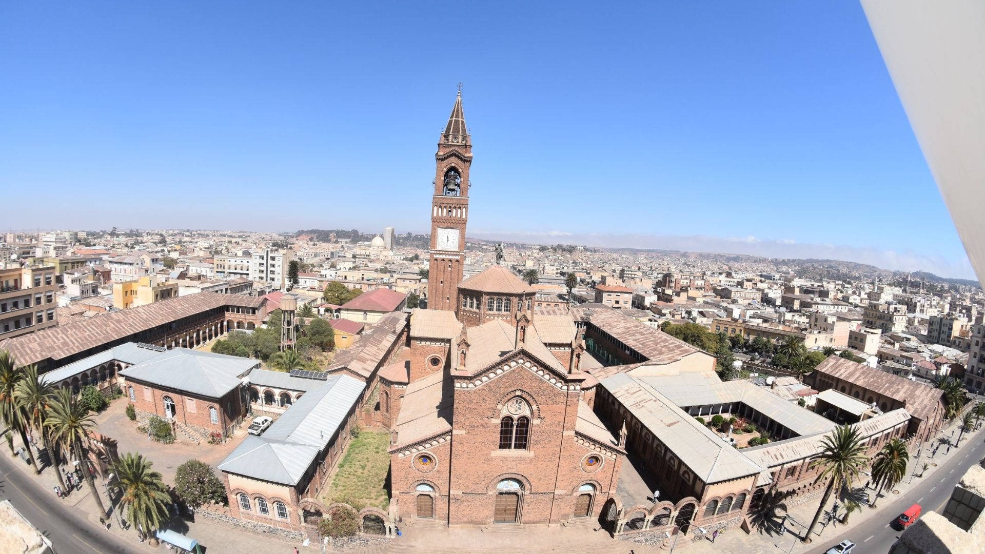 Eritrea 桌布 壁紙 — 歷史磚砌教堂的鳥瞰圖，高聳的鐘樓在晴朗藍天下主宰著陽光普照的地中海城市景觀。